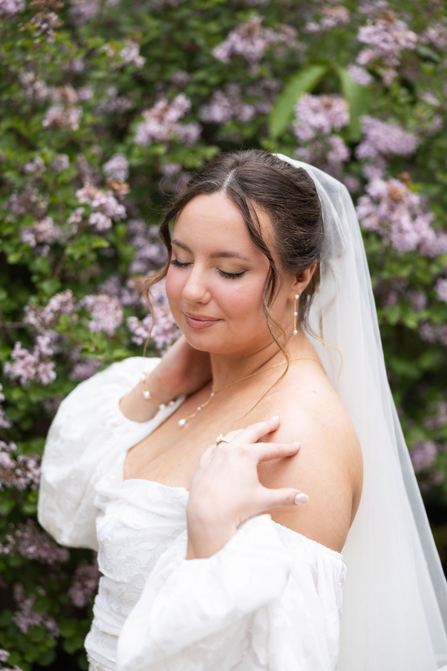 Bride in white off-shoulder gown and veil poses eyes closed before purple lilac bushes.