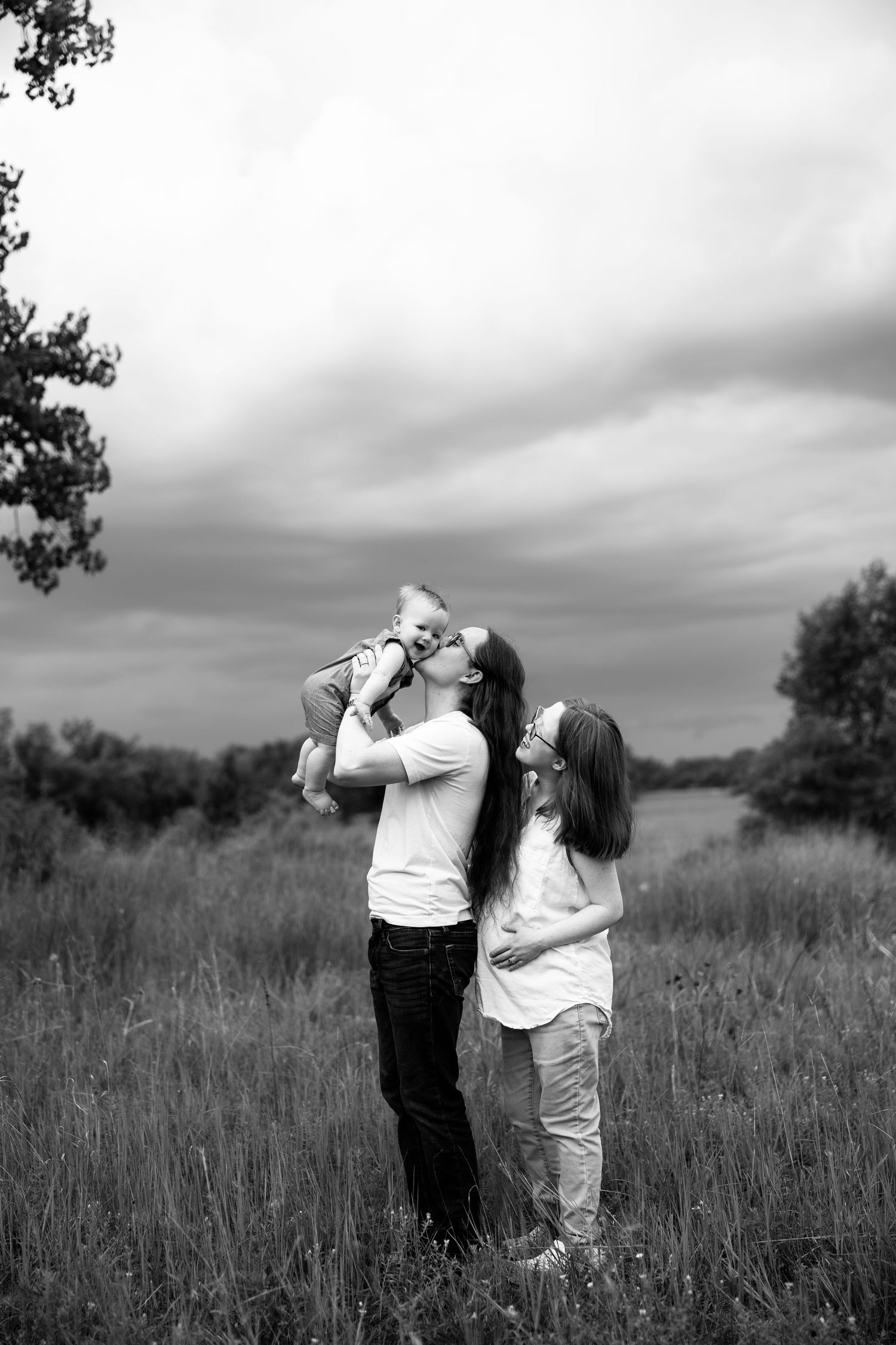 Black and white photo of parents kissing their baby in a field with dramatic cloudy sky.