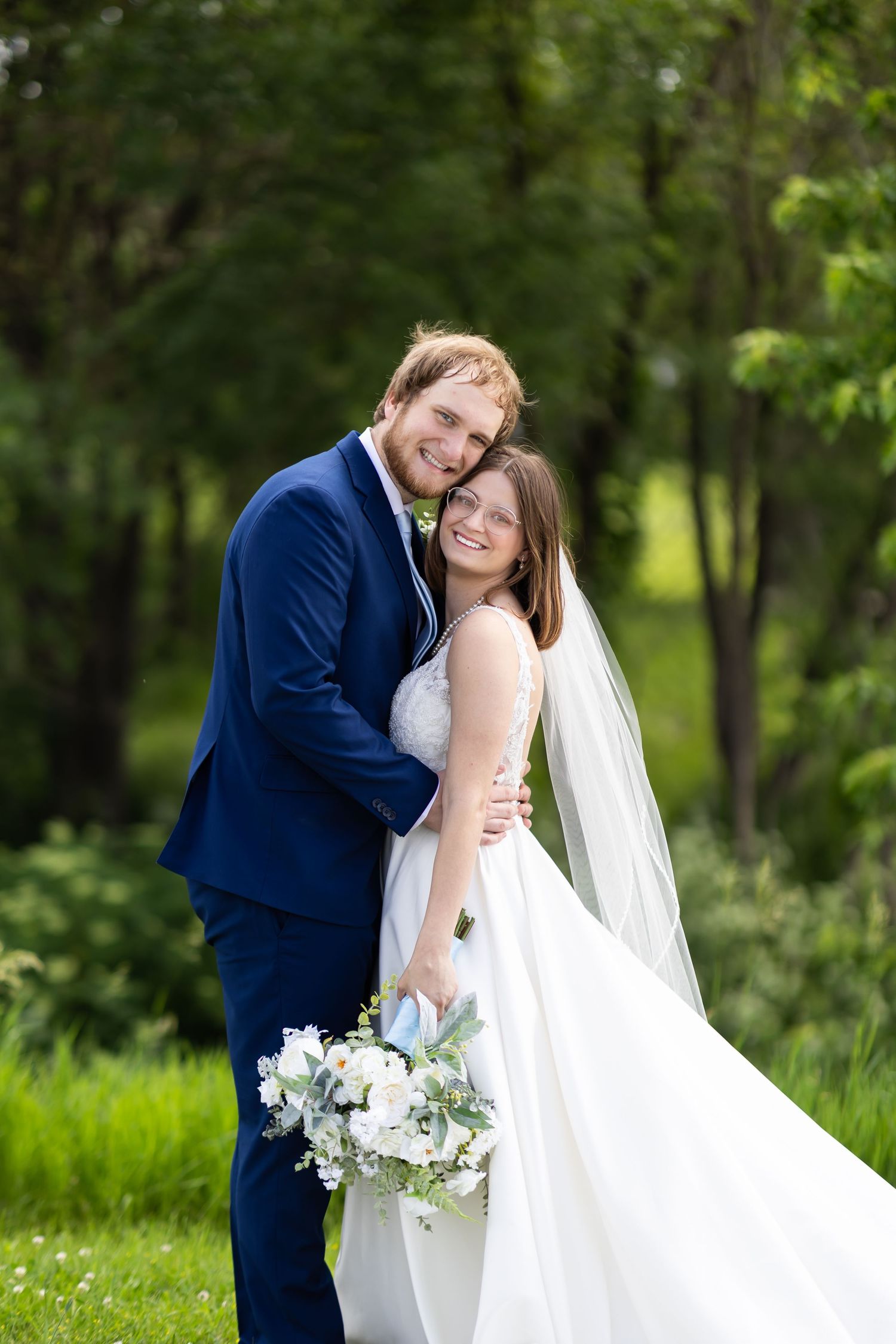 Newlyweds embrace in lush garden setting with white floral bouquet.