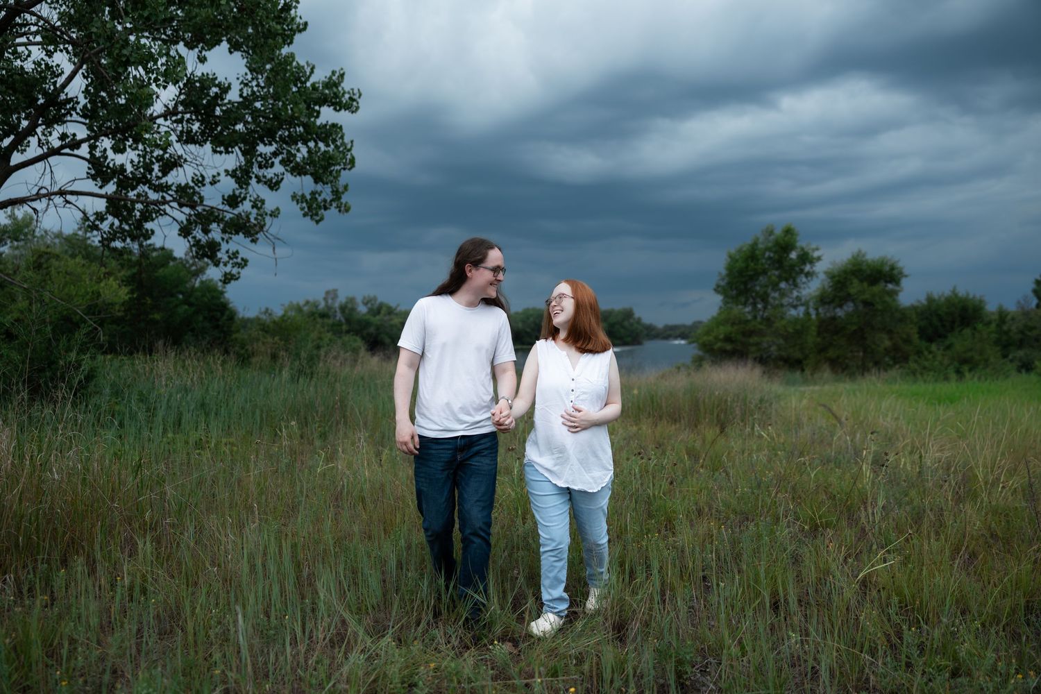 Couple in white shirts holding hands in a grassy field under dramatic stormy skies.