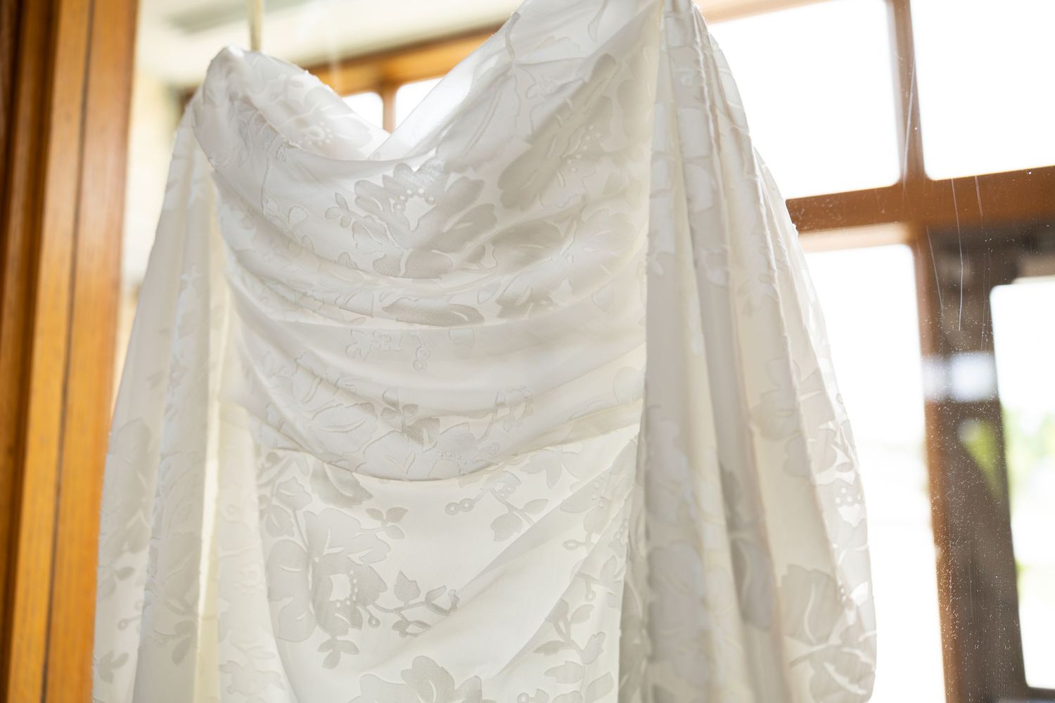 Elegant white lace wedding dress hanging by a window, showcasing intricate floral patterns and a sweetheart neckline.