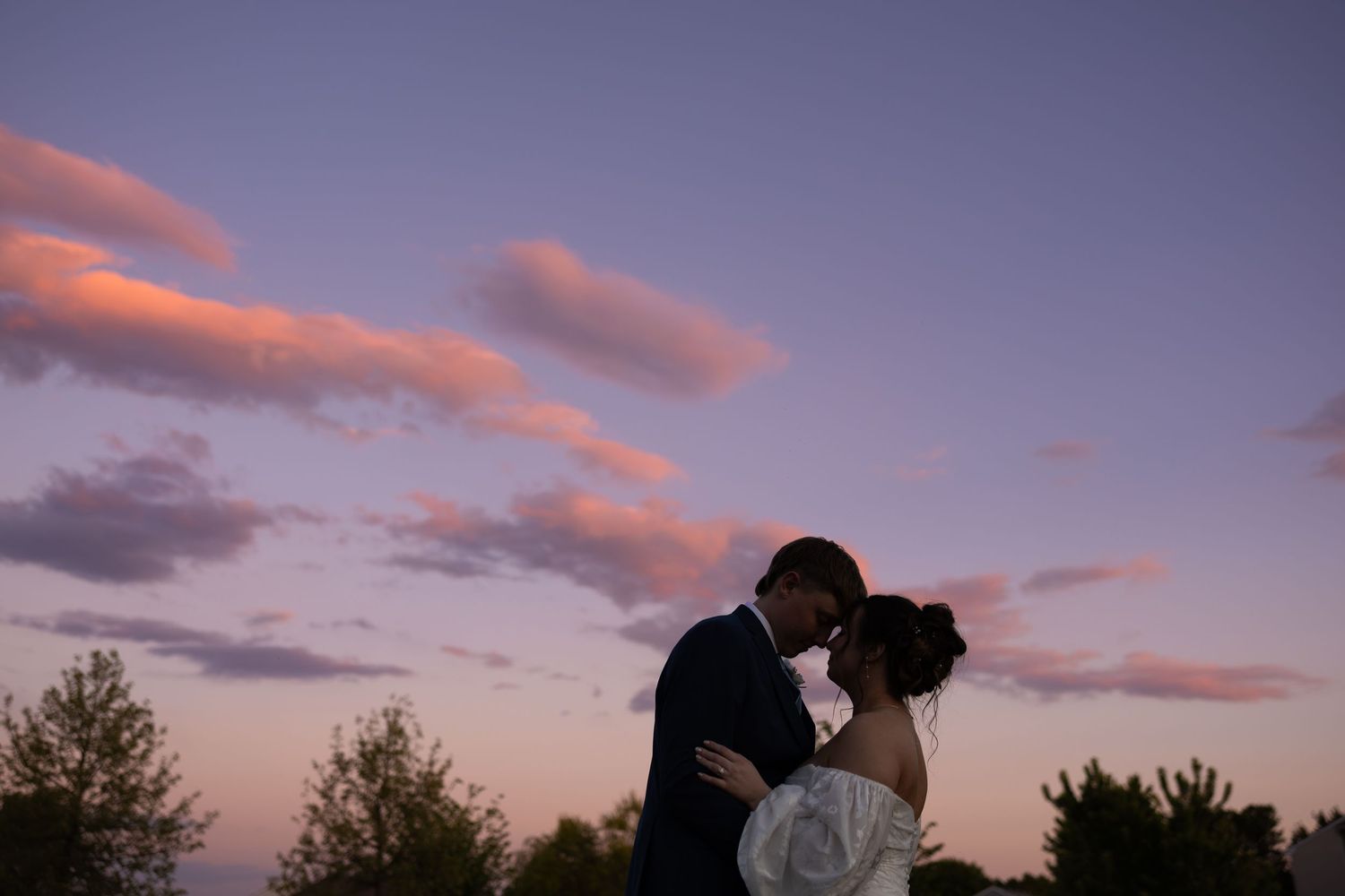 Bride and groom share a romantic kiss silhouetted against a stunning purple and pink sunset sky.