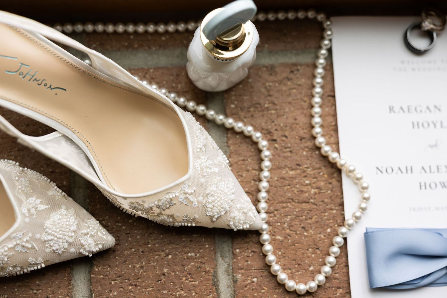 Elegant wedding flat lay featuring lace bridal heels, pearl necklace, perfume bottle, and wedding invitation on brick surface.