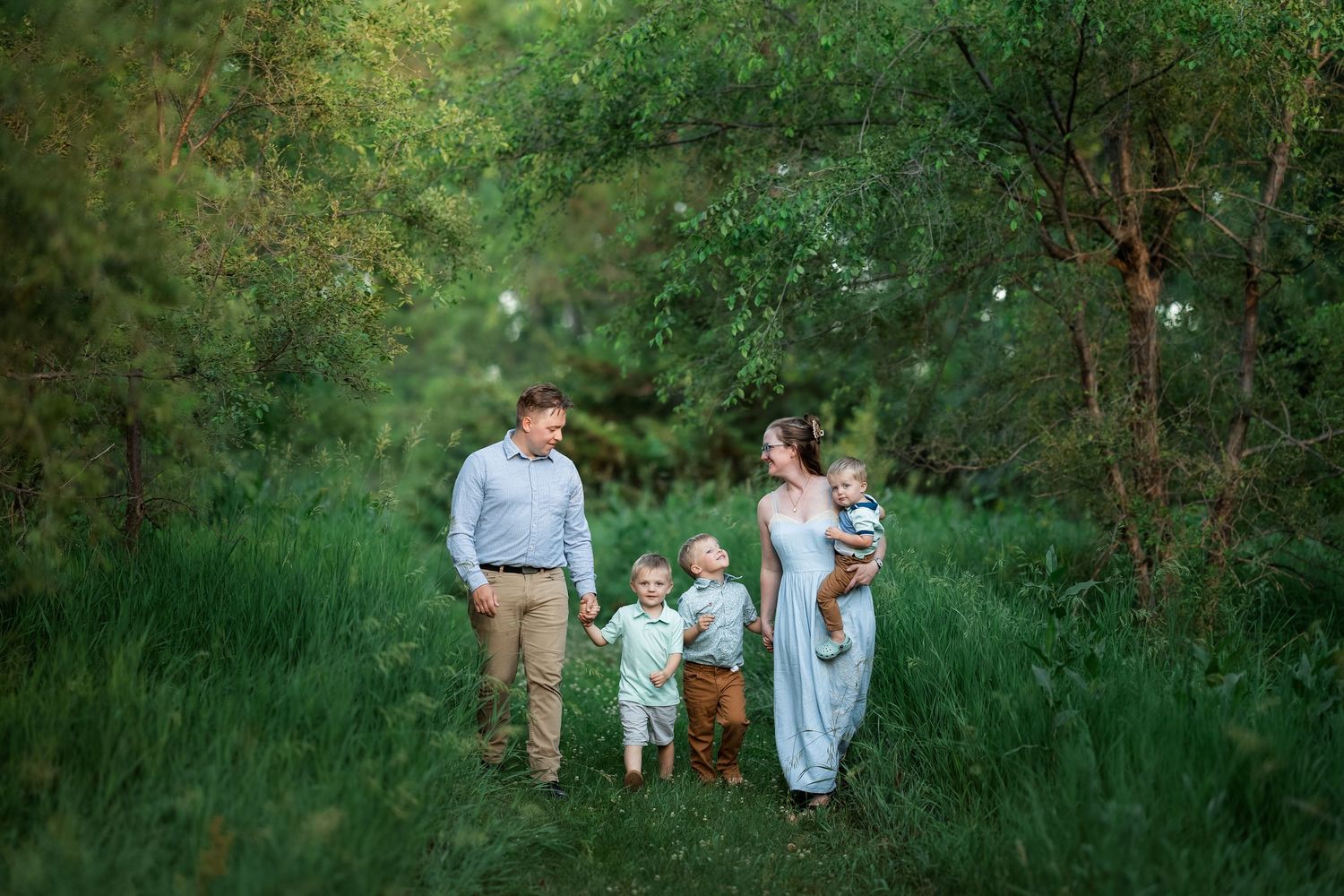 Family of four walks through lush green forest path, parents holding hands with toddlers on sunny day.