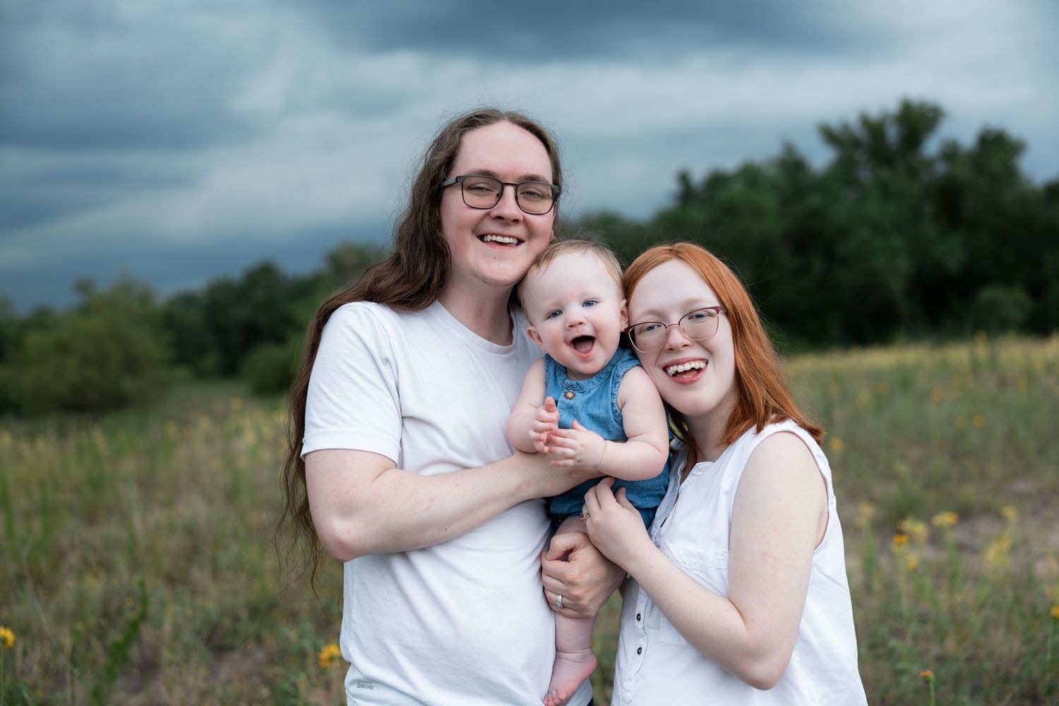 Two women holding a laughing baby in a field with stormy skies, wearing white outfits for a family portrait.