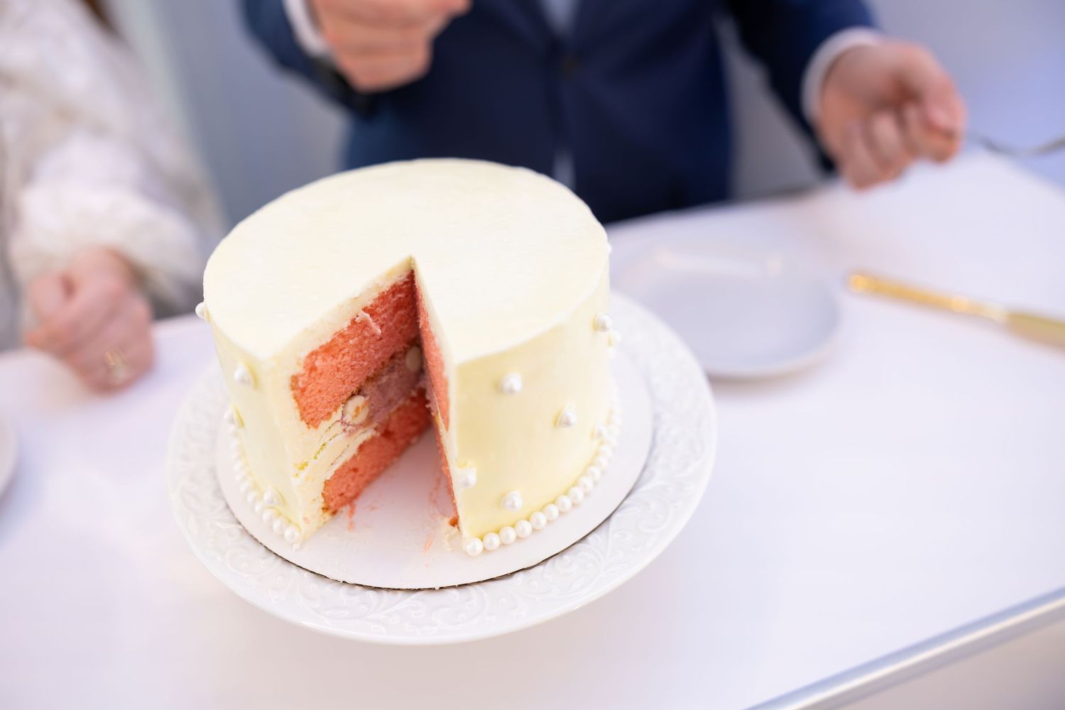 White frosted wedding cake with a slice removed revealing pink layers, on a white plate.
