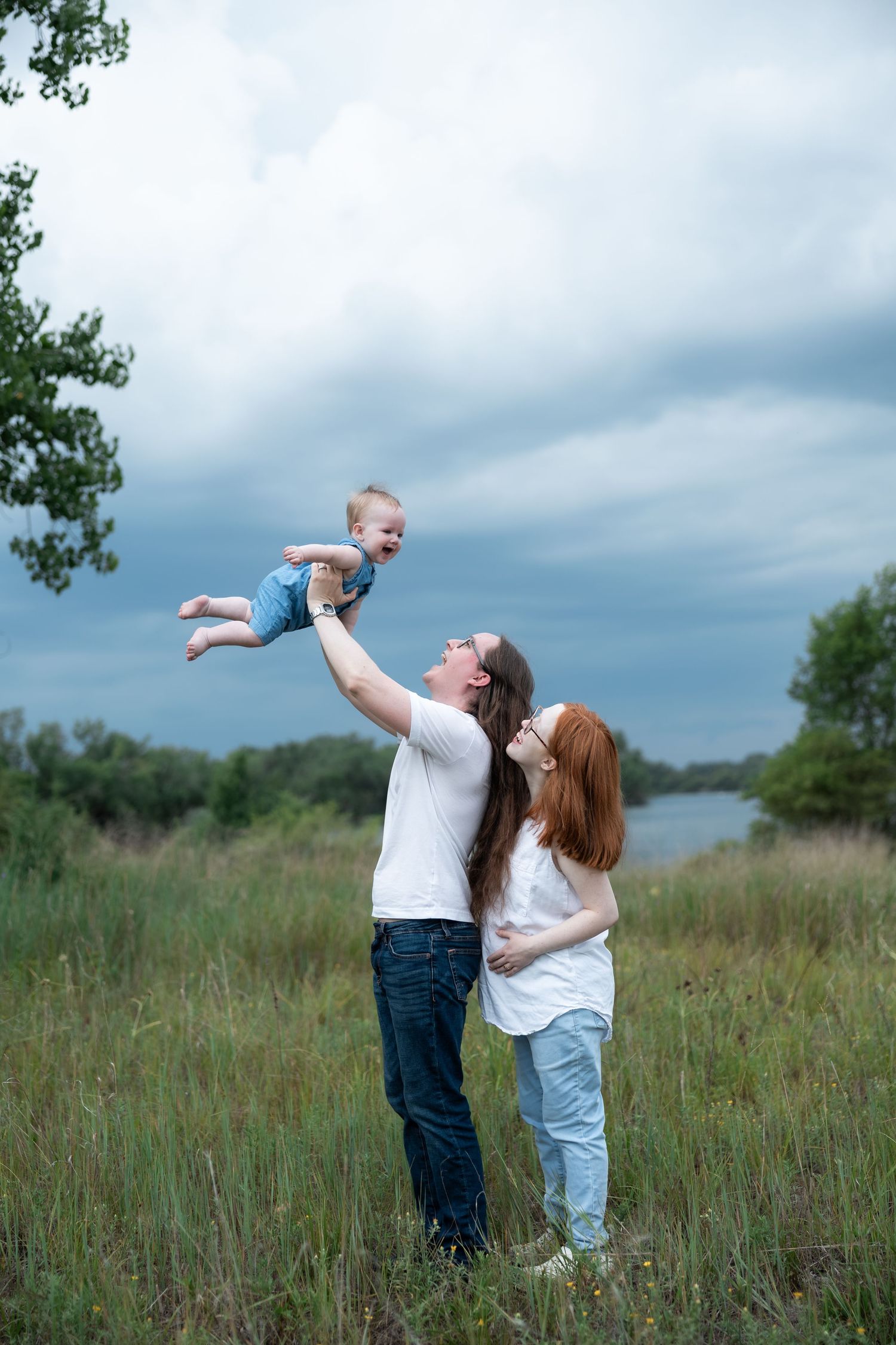 A happy family outdoors, father lifting laughing baby in the air while mother watches, standing in a green field.