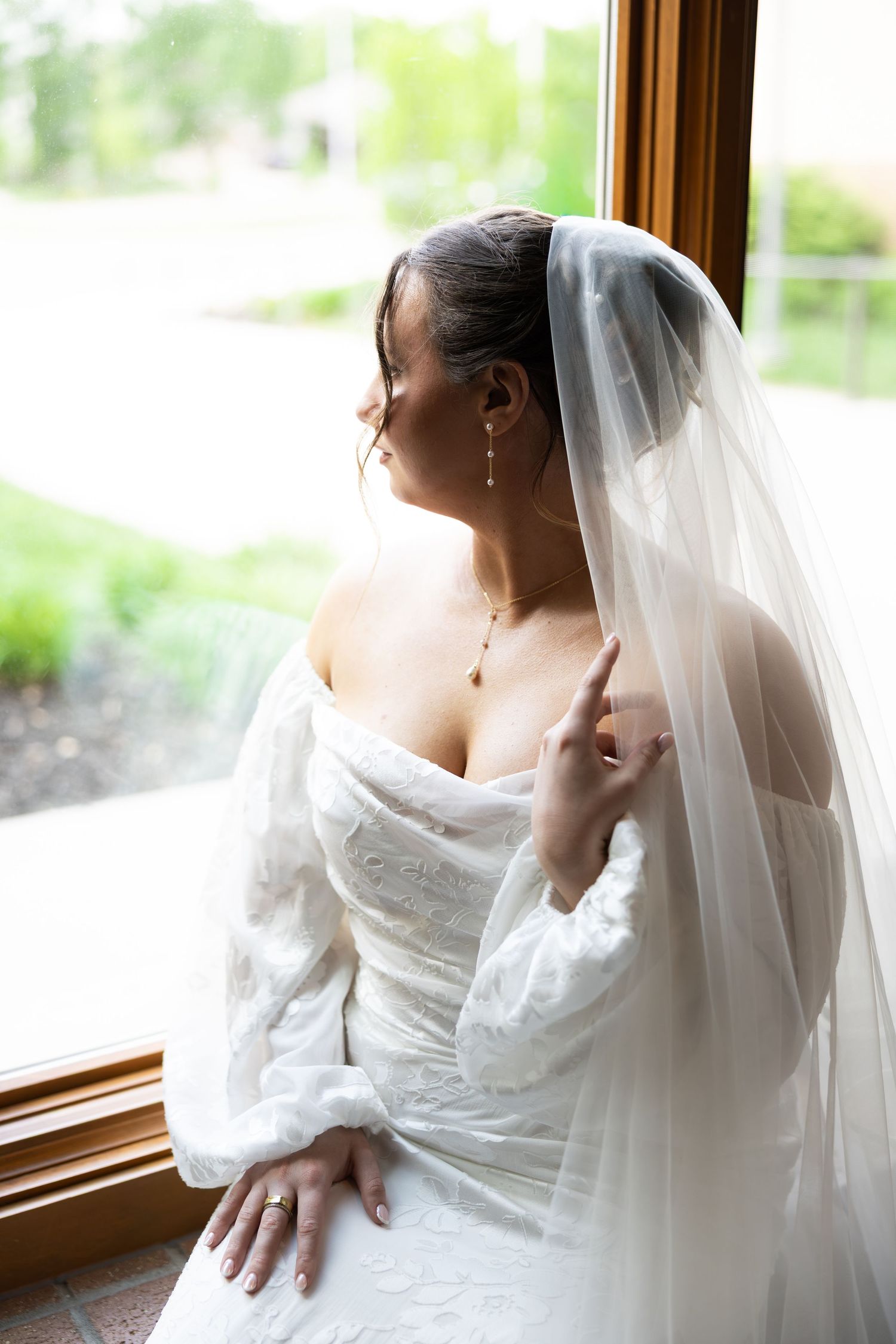 Bride in white gown and veil gazing out a window, wearing elegant drop earrings and necklace on her wedding day.