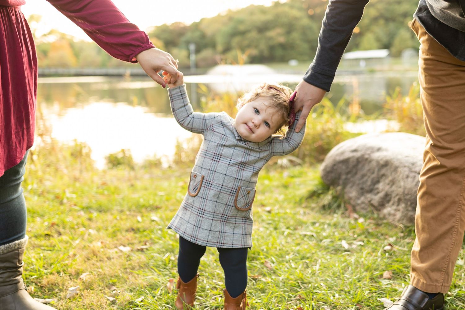Toddler in gray coat smiles while holding hands with parents outdoors by a lake at sunset.