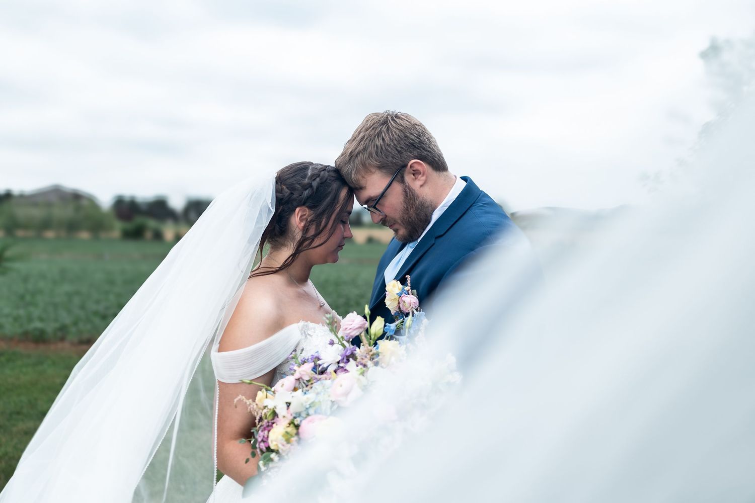 Bride and groom sharing an intimate moment, framed by a flowing veil, with a lush green field backdrop.