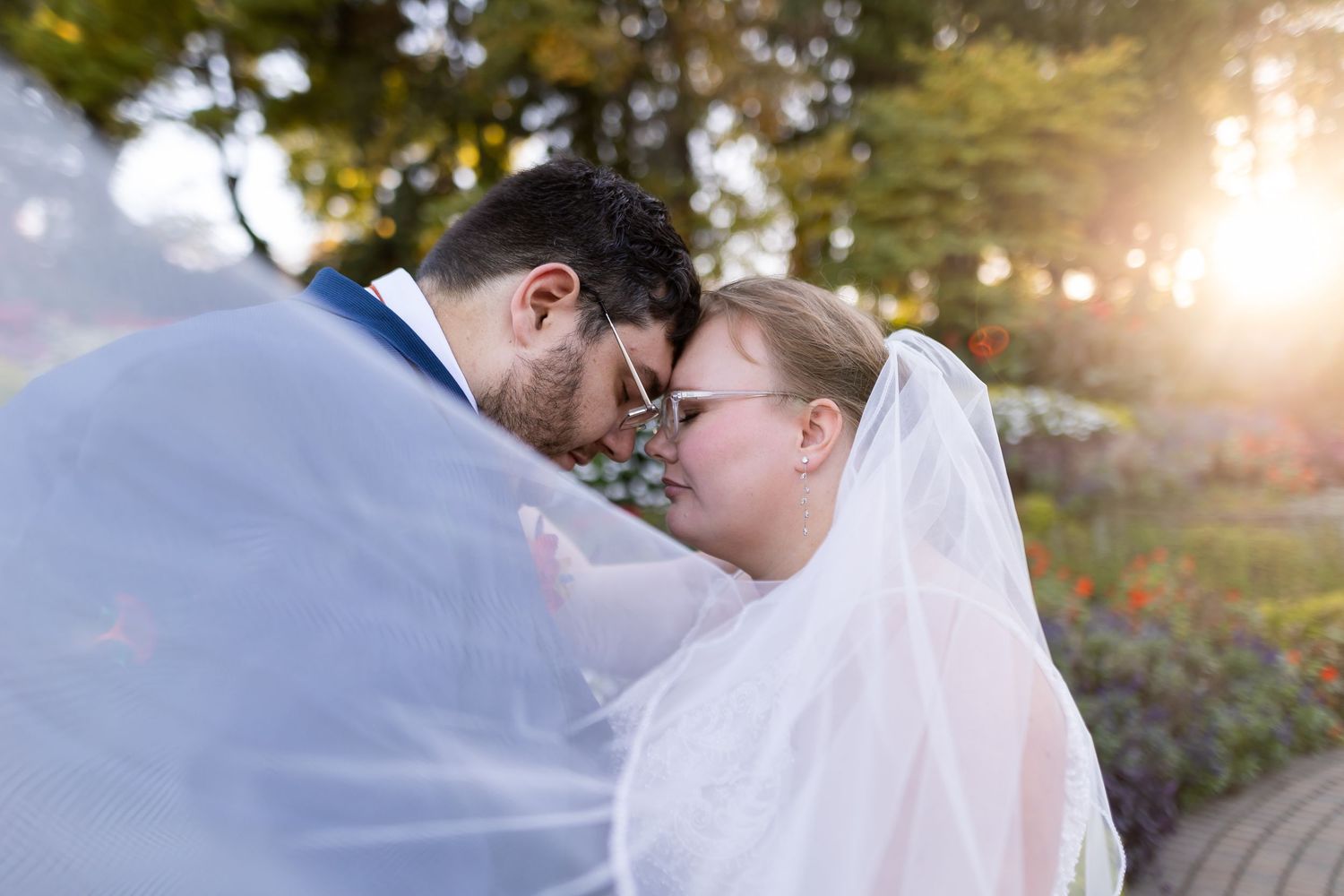 Bride and groom share an intimate forehead touch under a flowing veil, bathed in golden sunlight outdoors.