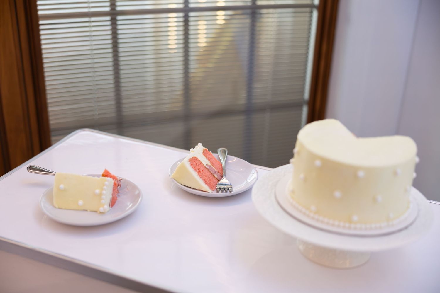 White frosted wedding cake on stand beside two plated slices revealing pink layers, displayed on white table near window.