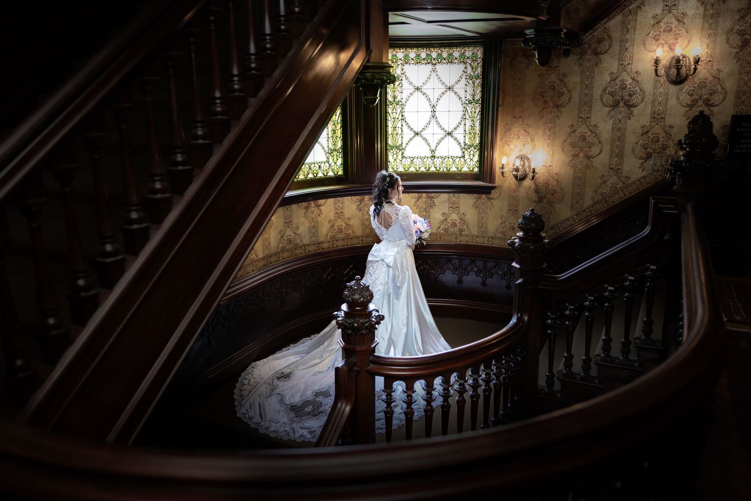 Bride in white wedding gown stands on ornate wooden staircase in elegant Victorian mansion with stained glass window.