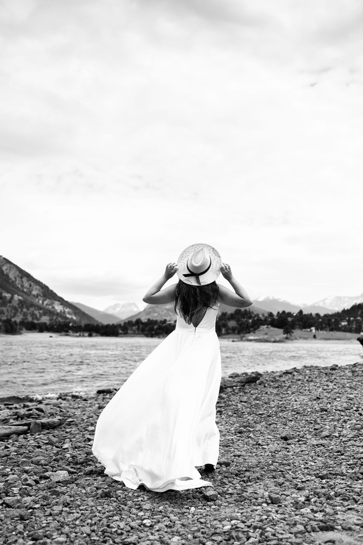 Woman in flowing white dress holds hat while standing by mountain lake in black and white photo.