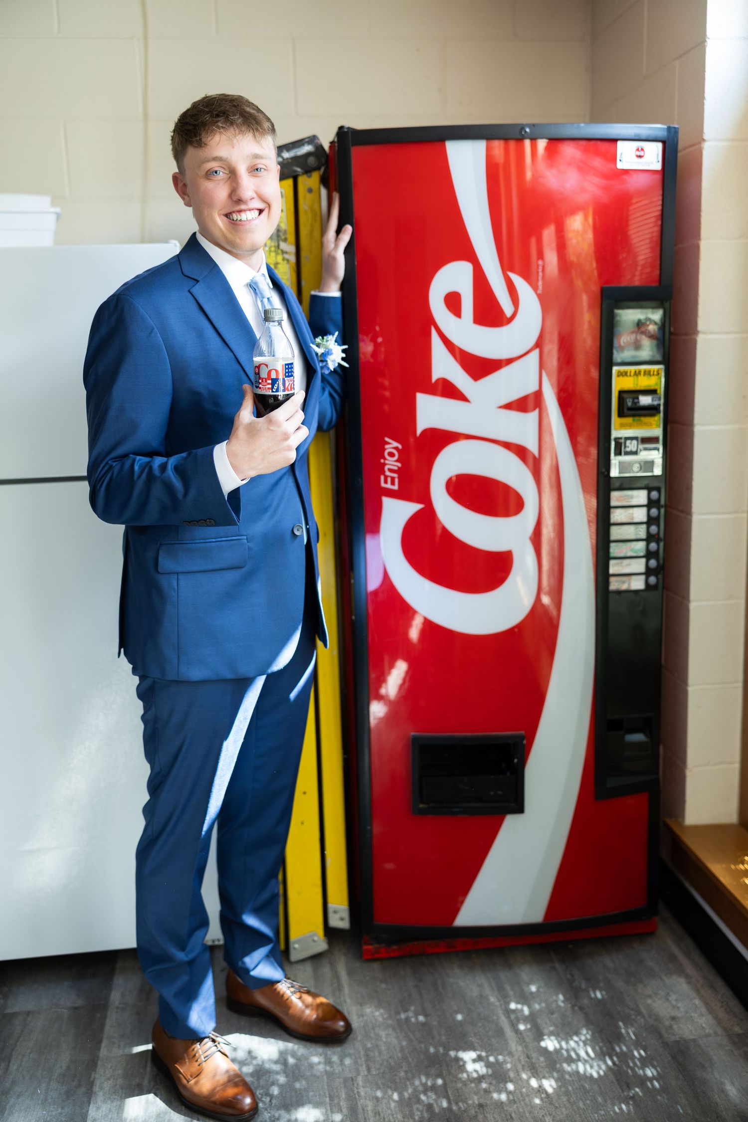 Young man in blue suit smiling beside an Enjoy Coke vending machine, holding a Coca-Cola bottle.