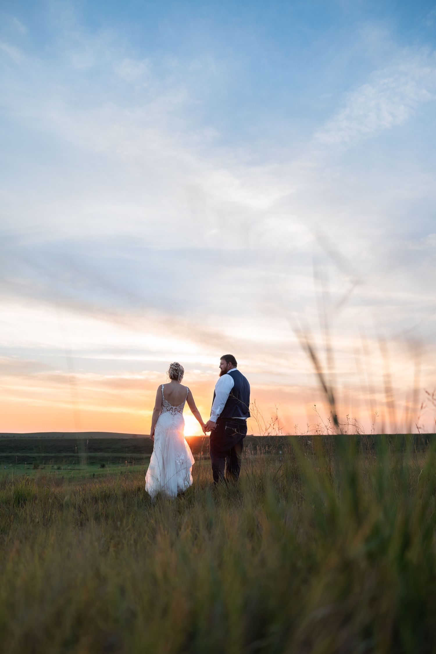 Bride and groom holding hands in a golden field at sunset, viewed from behind in wedding attire.