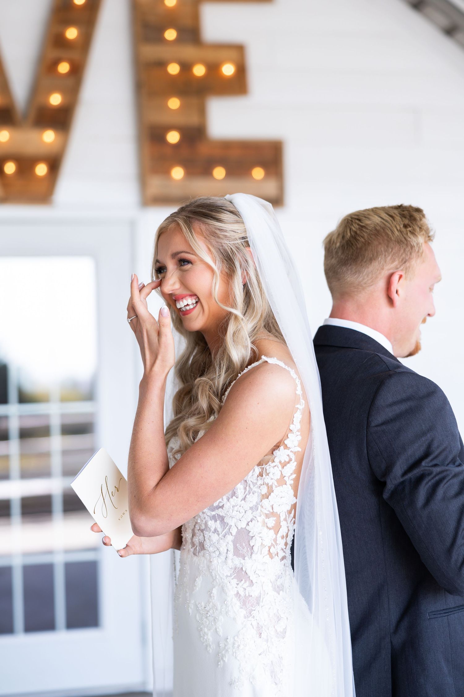 A beautiful moment captured during a wedding first look as a bride in lace dress smiles with joy next to her groom.