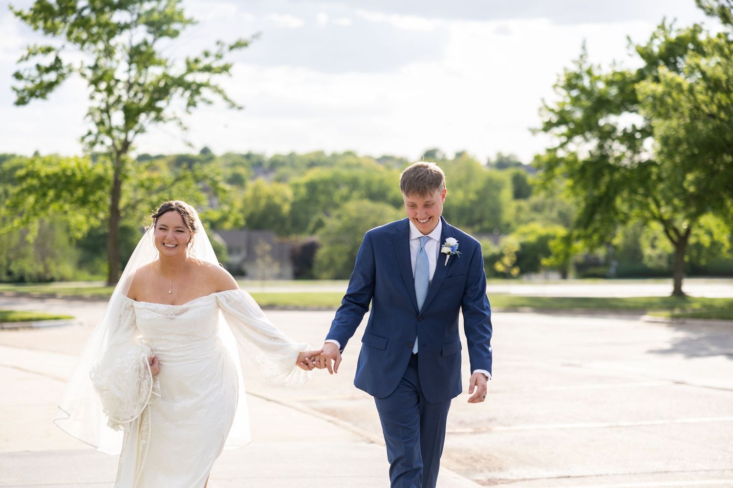 A bride in a white gown and groom in a navy suit hold hands while walking outdoors on their wedding day.