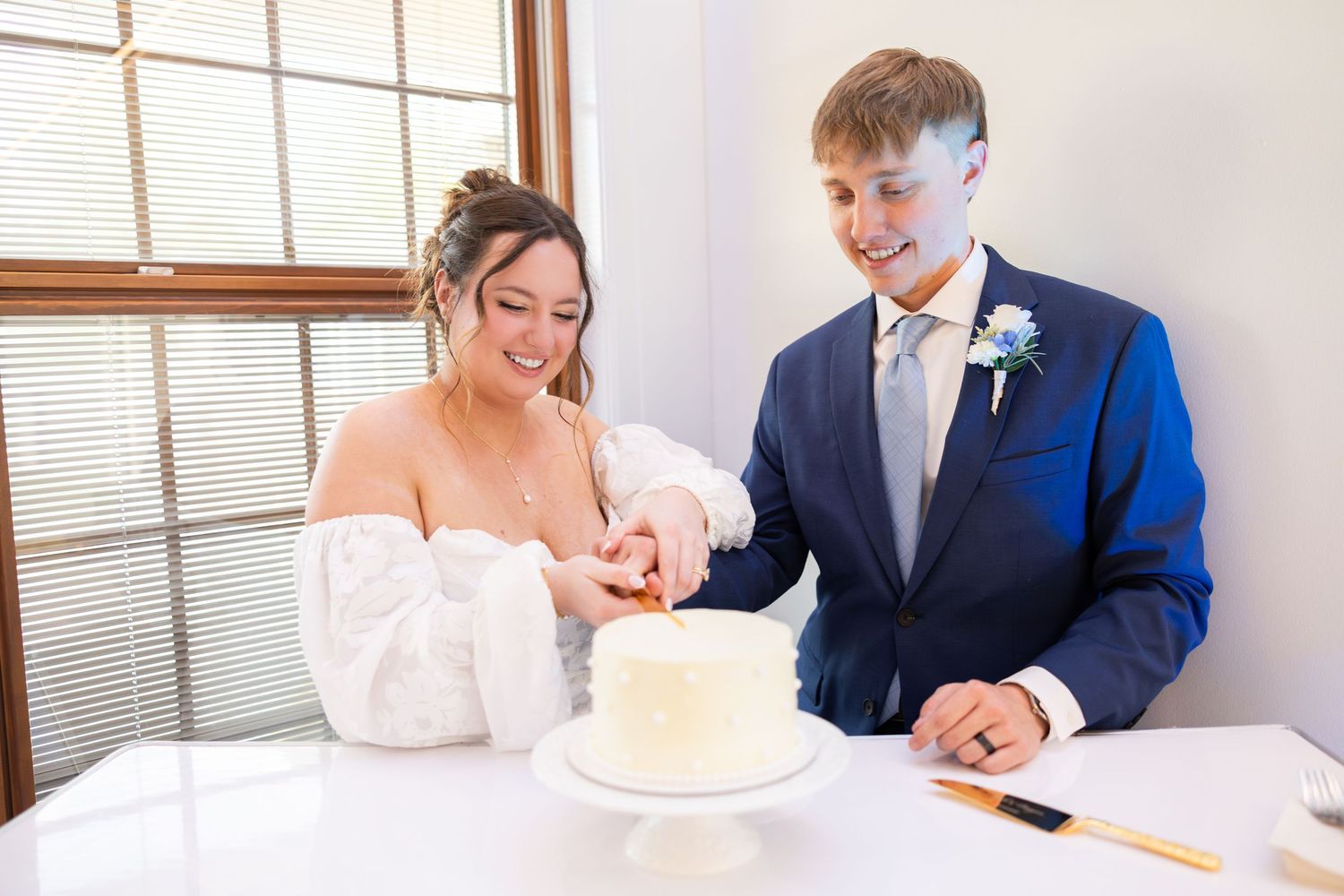 Bride and groom cutting white wedding cake together, groom in navy suit with boutonniere, bride in white dress.