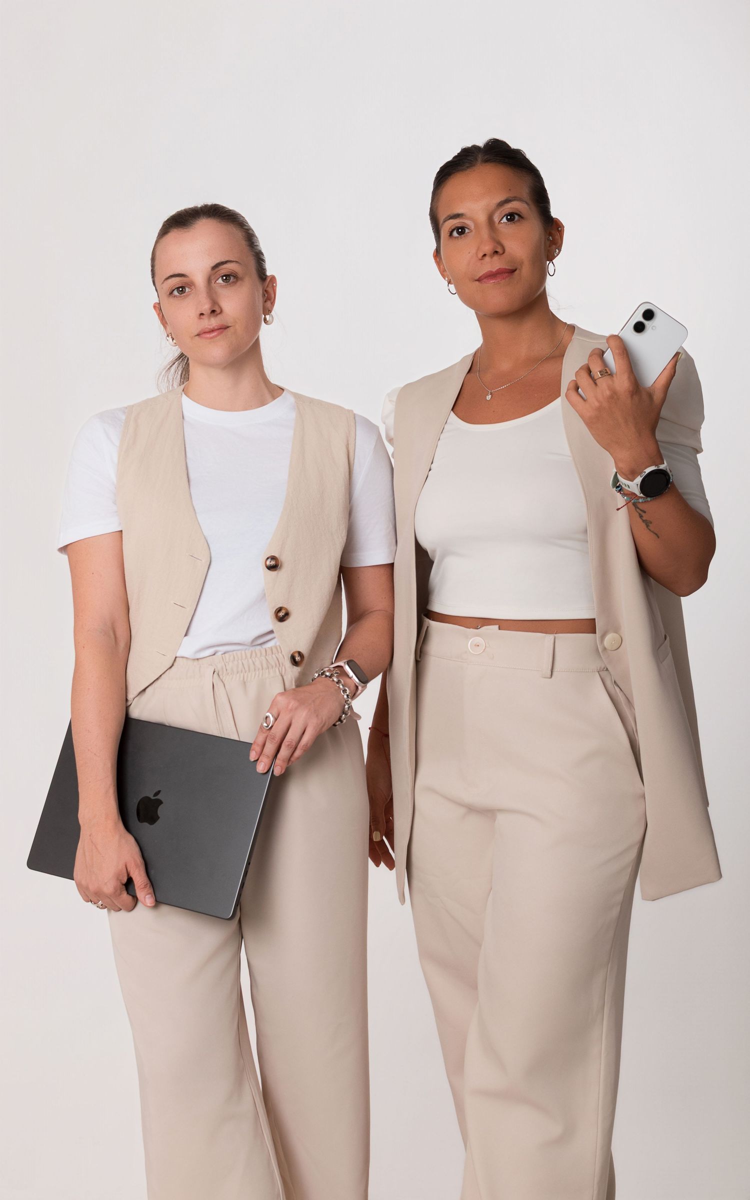 Retrato grupal corporativo en equipo, de dos mujeres con outfits en tonos beige y blanco, una sosteniendo una notebook y la otra un celular.