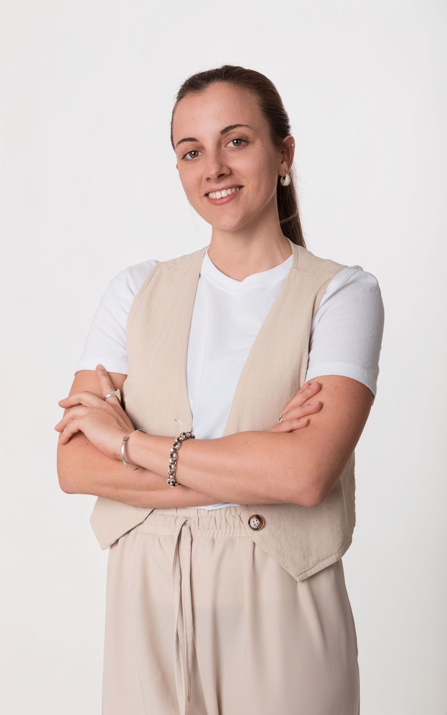 Retrato profesional individual de mujer con chaleco beige y remera blanca, posando con los brazos cruzados sobre fondo blanco.