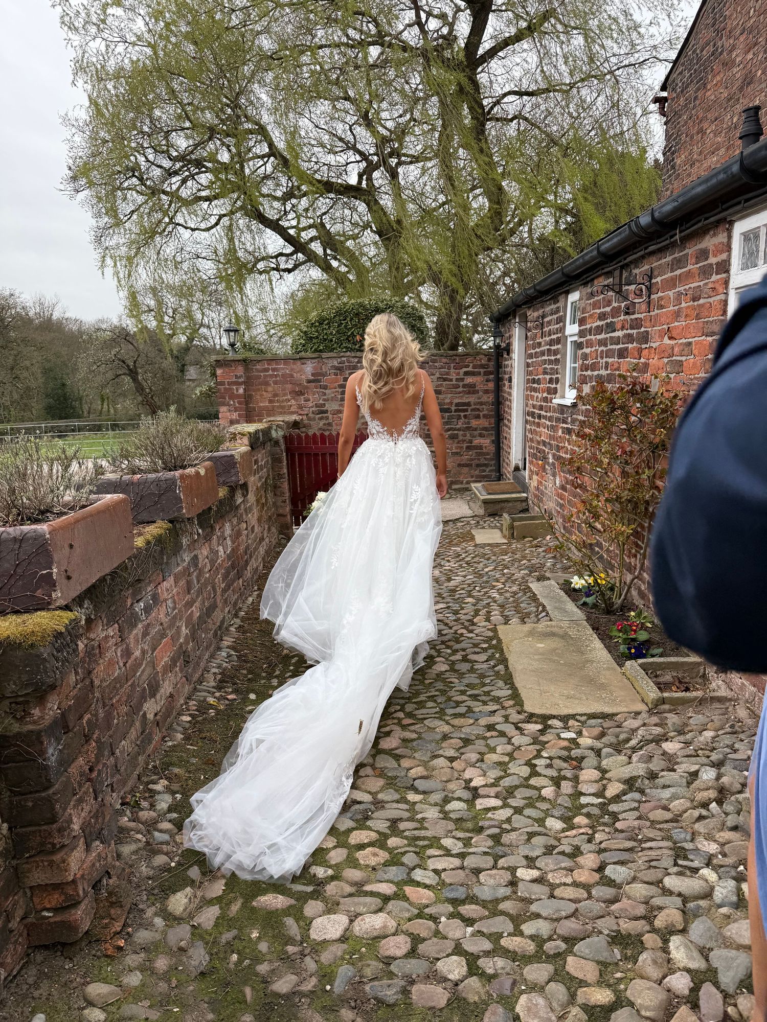 Bride in flowing white gown walks along cobblestone path beside brick building.