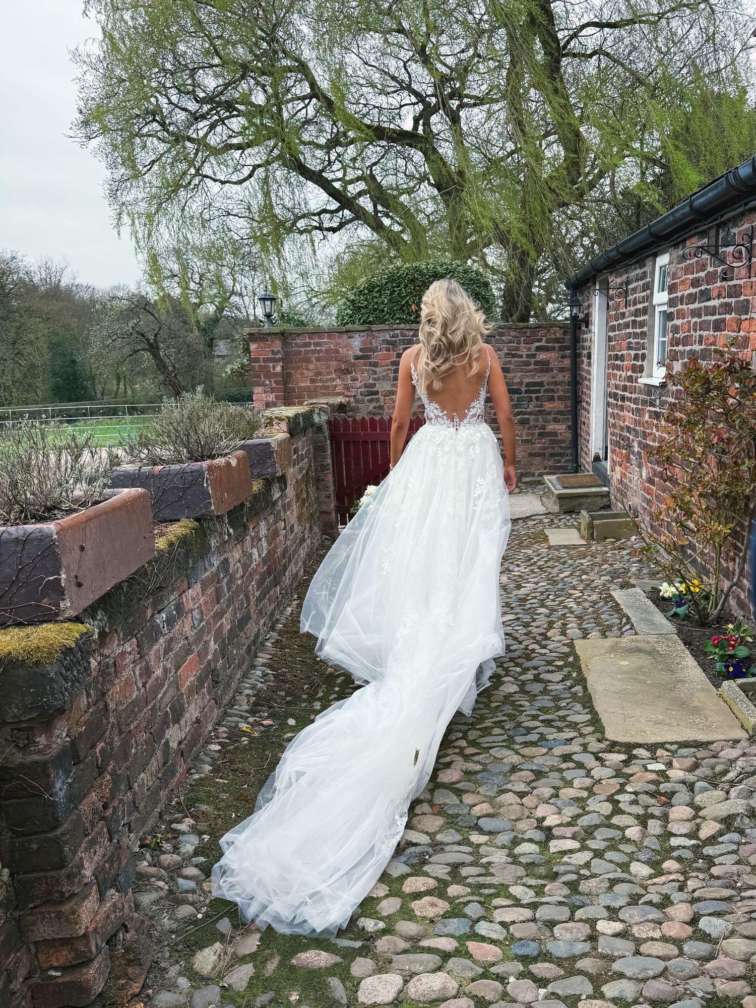 Bride in lace wedding dress walks along historic cobblestone path near brick wall.