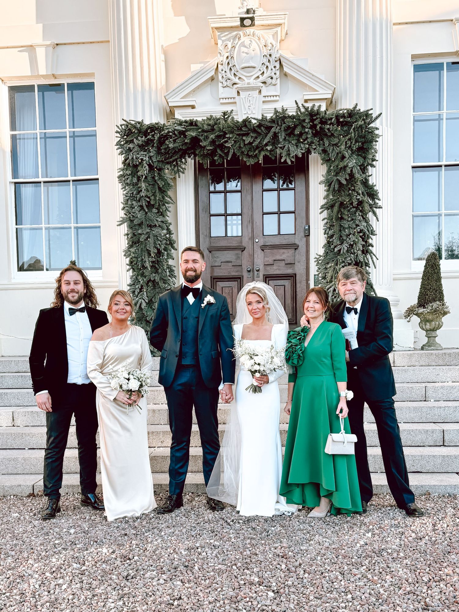 Formal wedding party lineup against white pillared building with evergreen decor.
