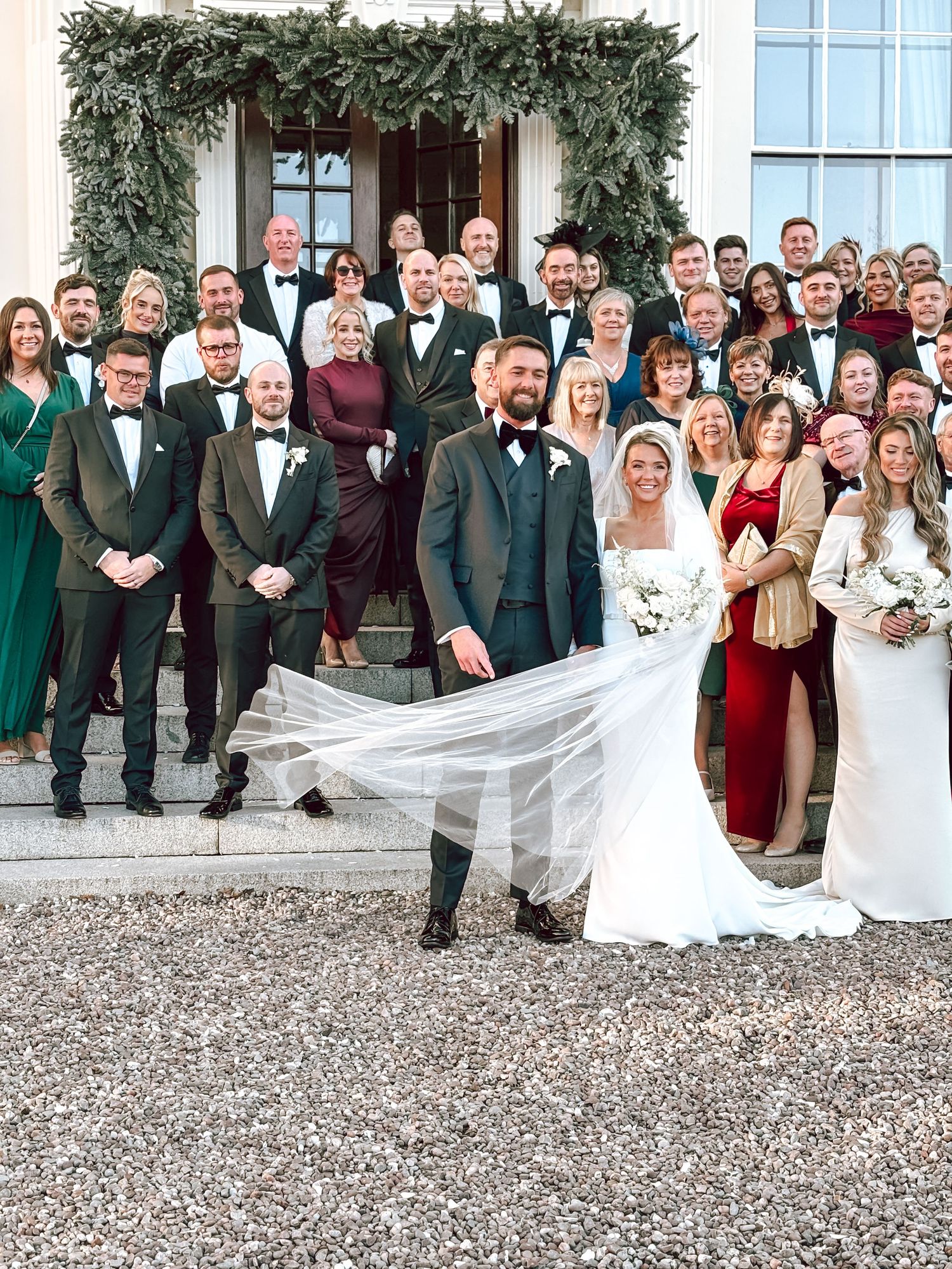 Large wedding party group photo outdoors with greenery and formal attire.