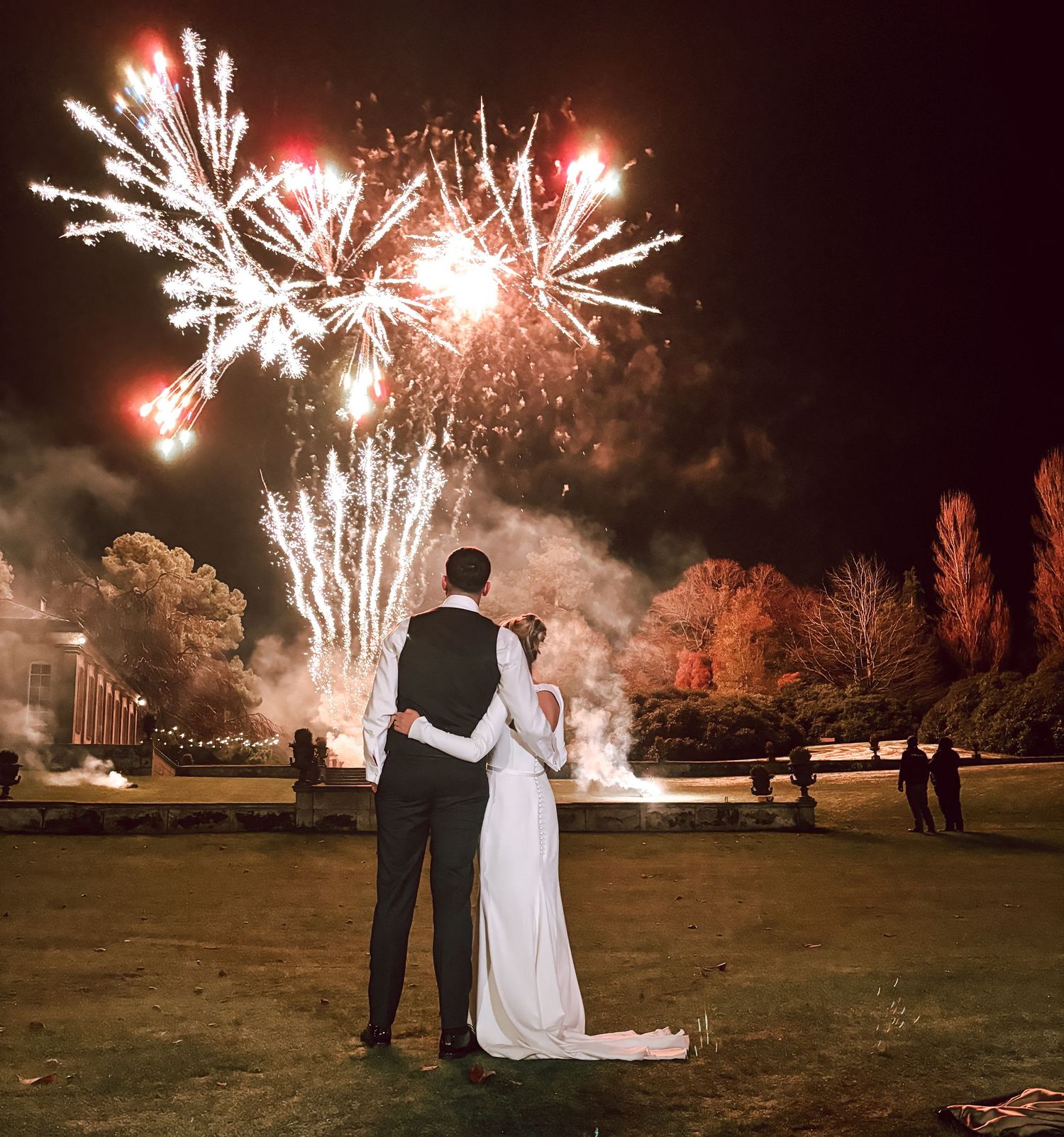 Silhouettes of a couple watching bright red and white fireworks display in a nighttime outdoor setting.