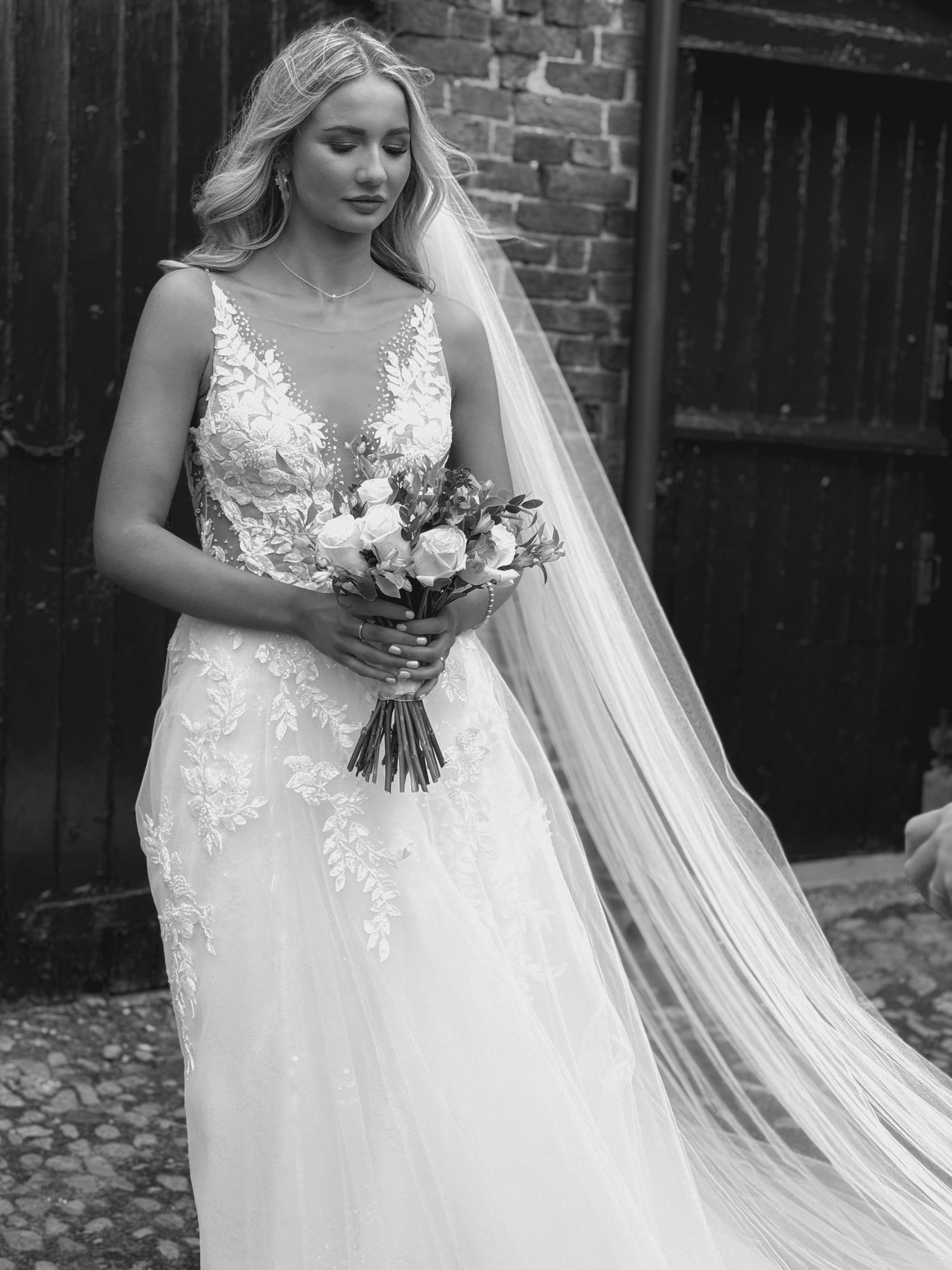 Bride in detailed lace gown holds white and pink bouquet against brick backdrop.