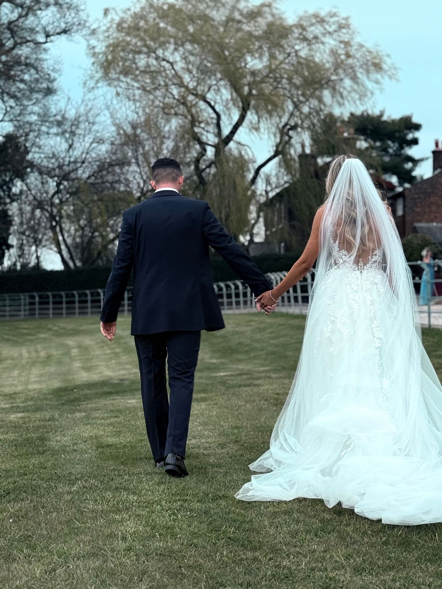 Wedding couple walking together across grassy field with flowing bridal gown.