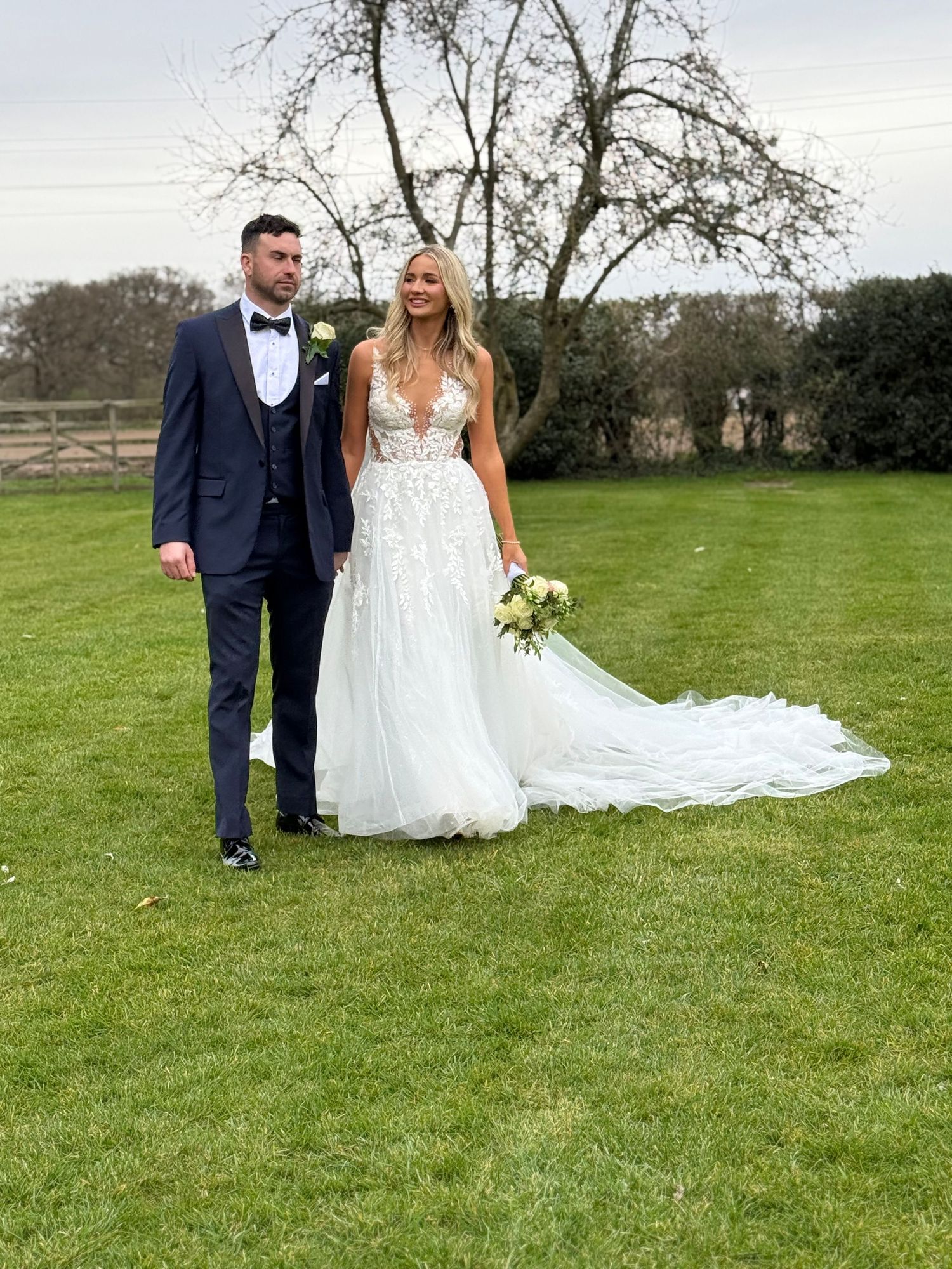 Newlyweds pose together on green lawn with flowing white wedding dress train.