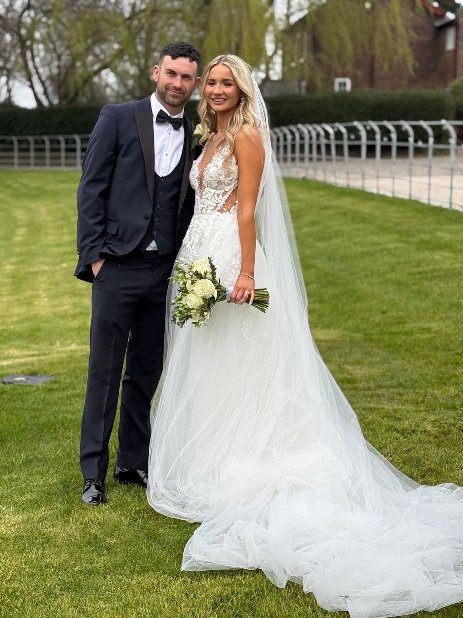 Wedding couple poses together on grass with bride's dramatic train flowing behind.