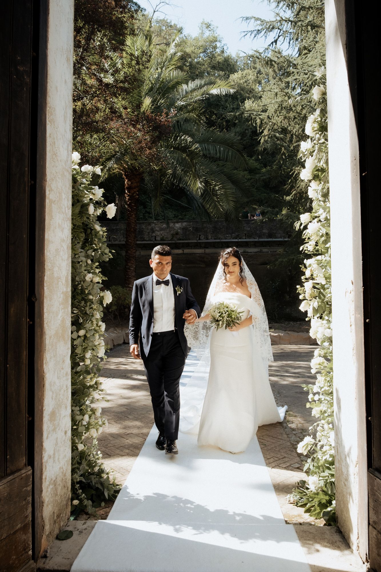 Wedding ceremony setup with white floral arch and aisle runner in an outdoor garden setting.