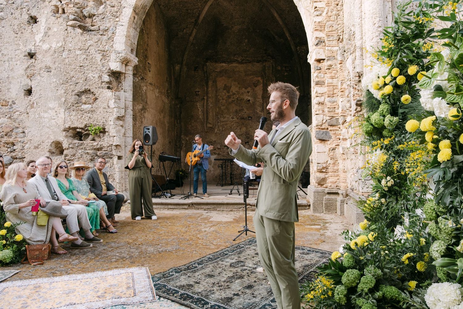 Wedding ceremony in a rustic stone courtyard with yellow flowers and greenery decorating ancient walls.