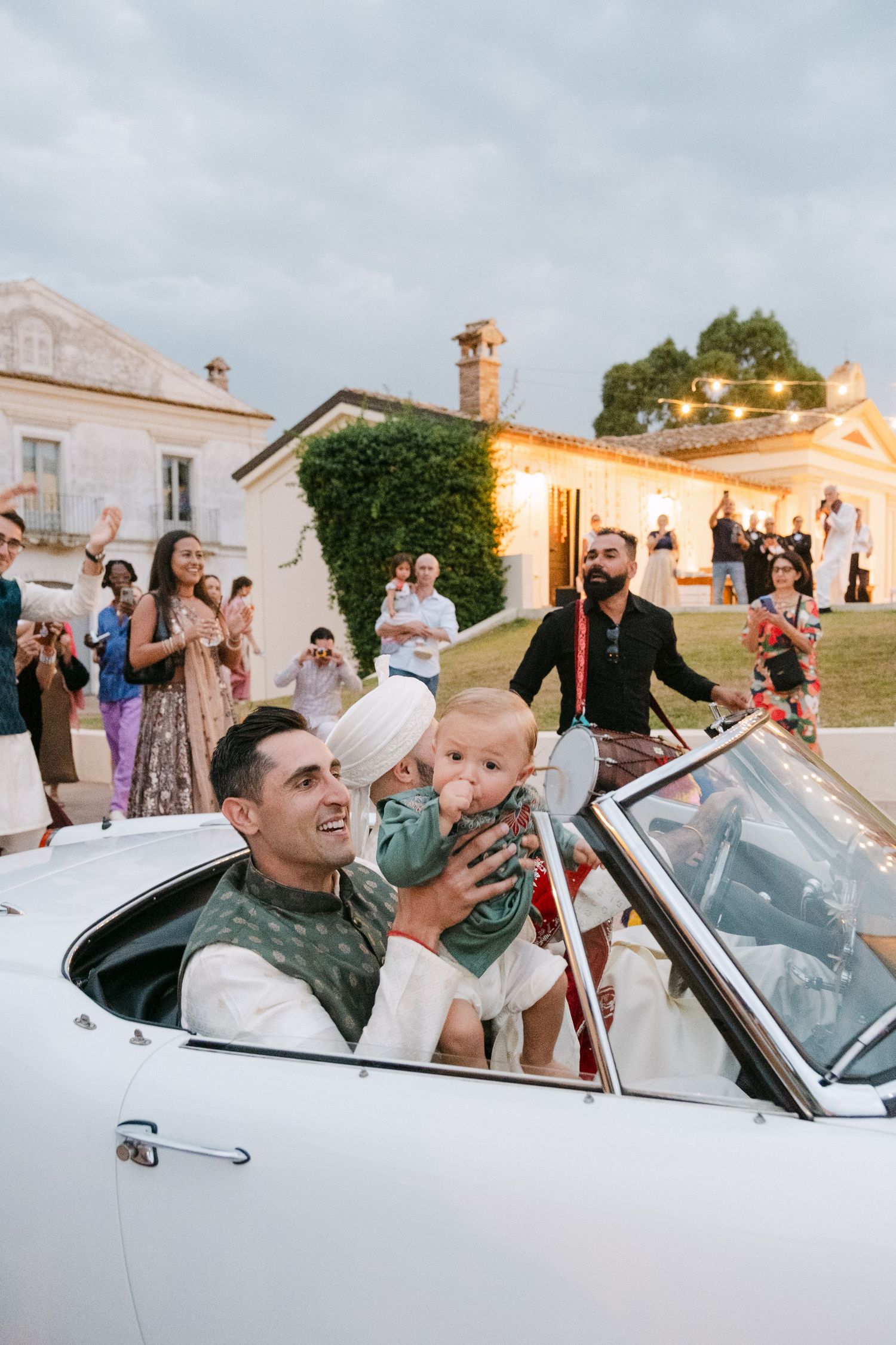 A classic white convertible car carries passengers during an outdoor celebration at a villa.