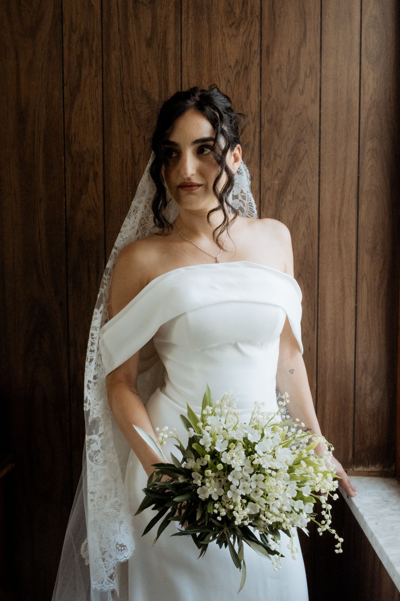 A bride in a strapless white wedding gown holds a delicate bouquet of white flowers and greenery.