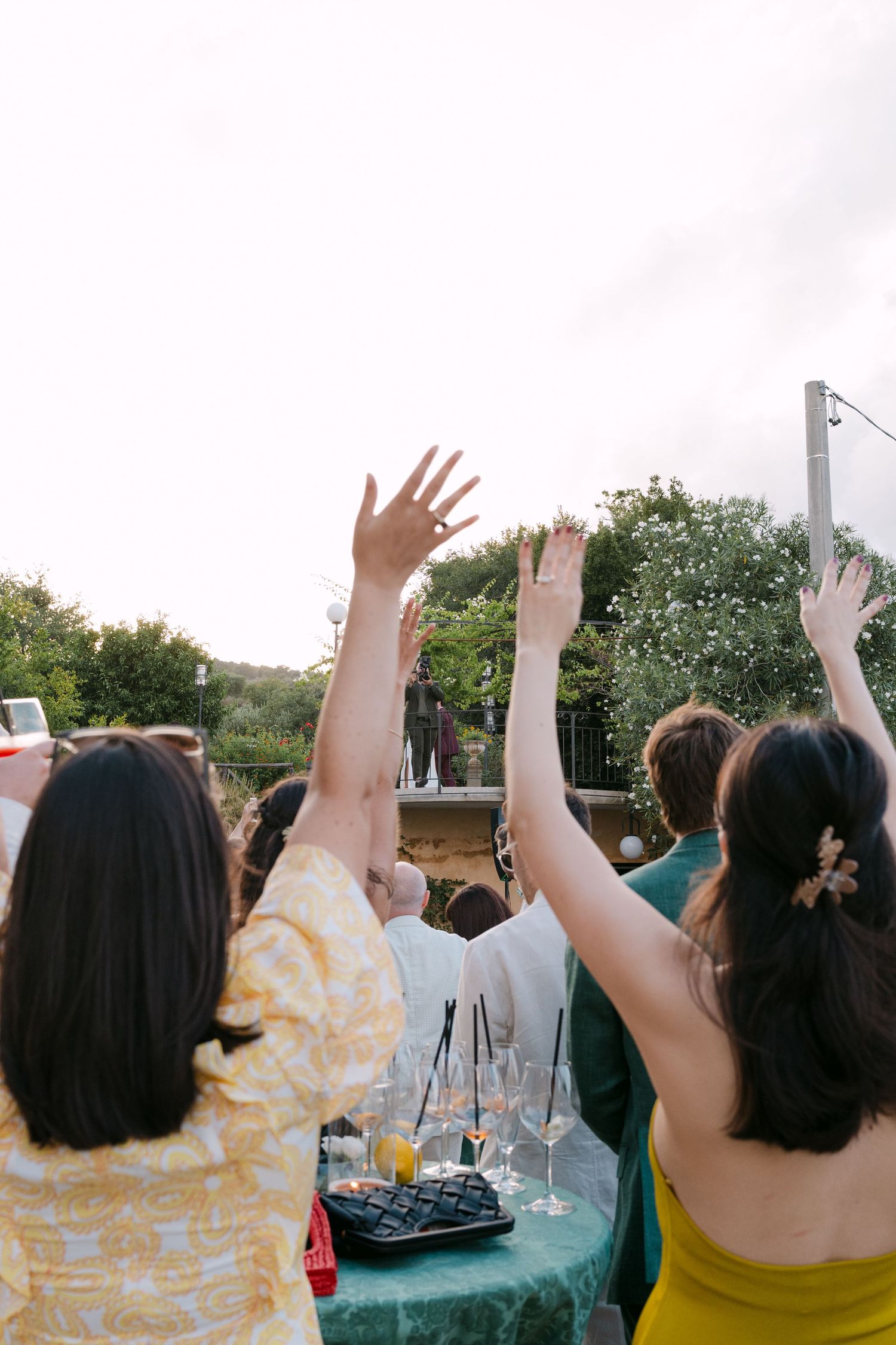 Wedding guests raise their hands in celebration against a sunset sky.