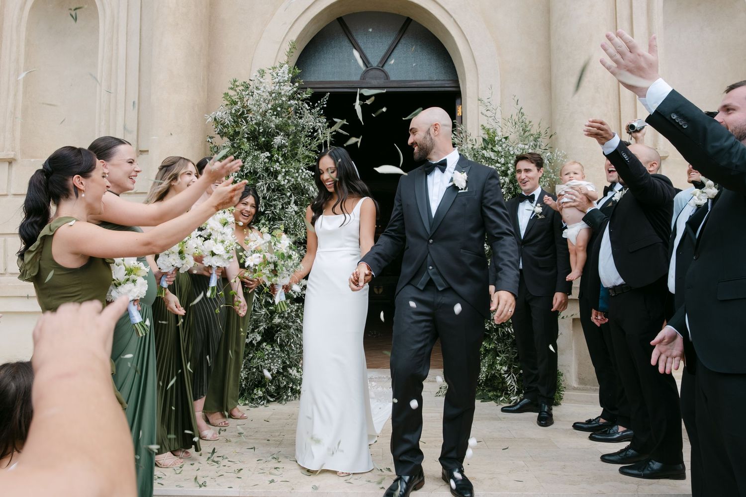 Wedding party celebrates with joy outside a church entrance with bridesmaids in olive green dresses and floral decor.