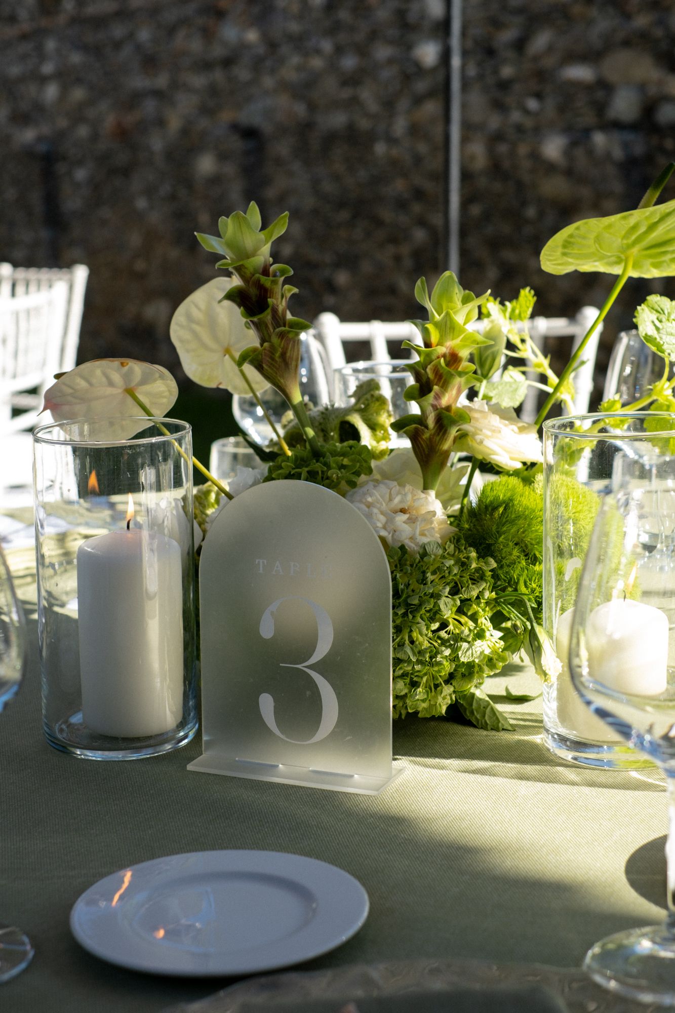 Elegant wedding table centerpiece with frosted glass table number, white candles and fresh greenery arrangements.