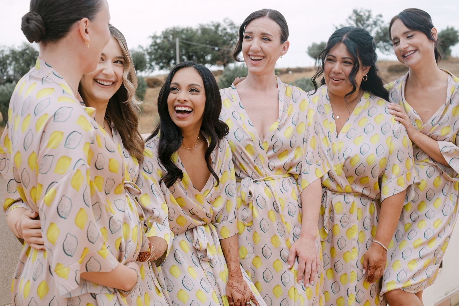 Bridesmaids wearing floral yellow and white robes laugh together during pre-wedding preparations.