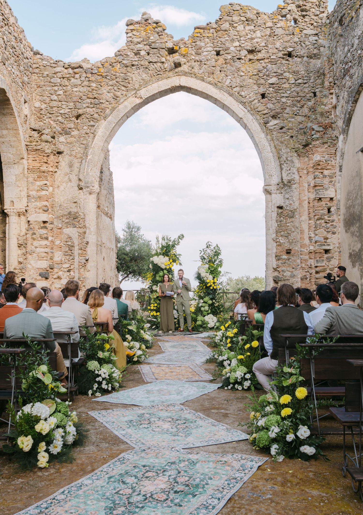 A wedding ceremony setup in ancient stone ruins with floral arrangements and a vintage rug aisle runner.