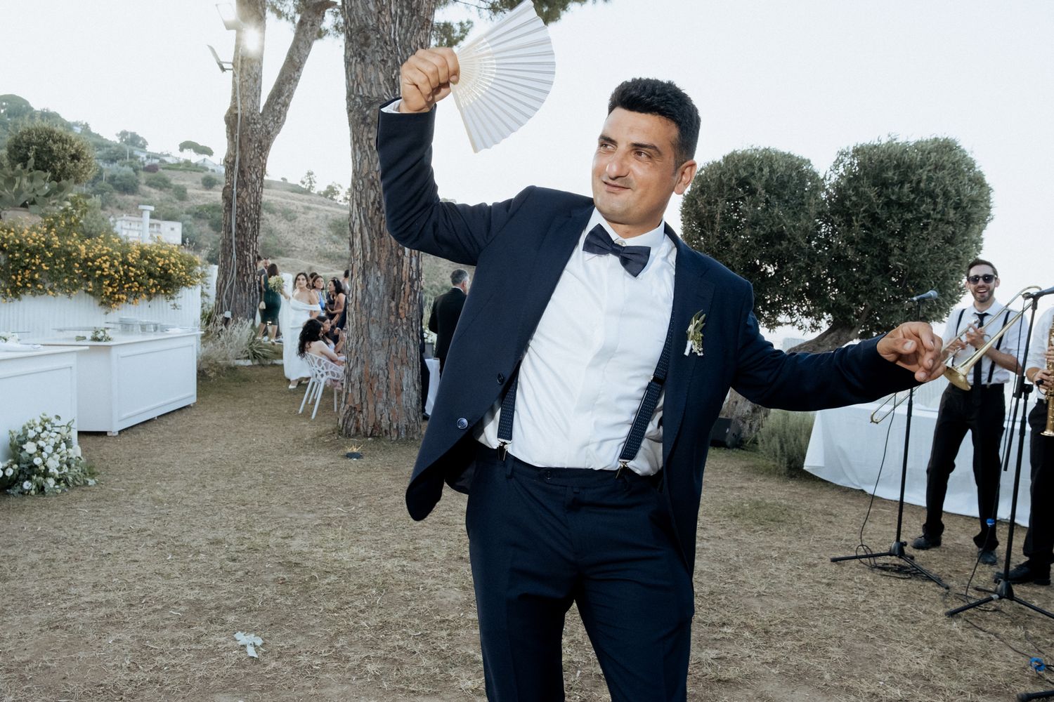 A person in a navy suit dances with a white fan at an outdoor wedding reception.