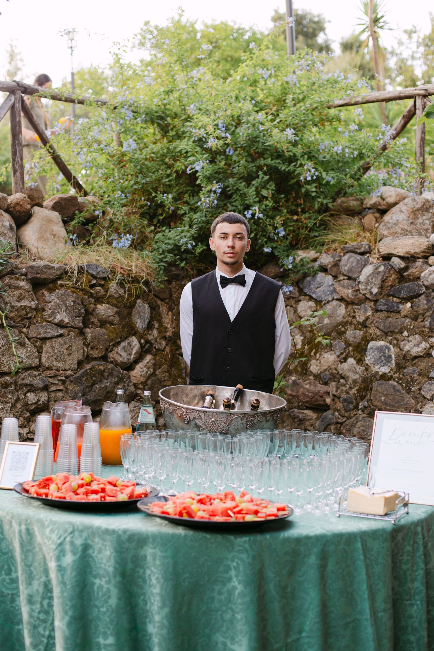 Catering table with watermelon plates and drinks set up against a stone wall outdoors.