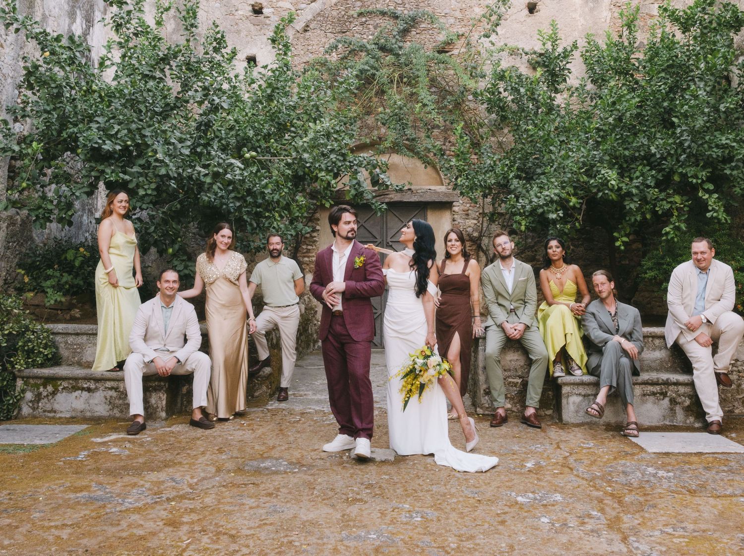 Wedding party in neutral and burgundy attire poses together in front of olive trees in an outdoor garden setting.