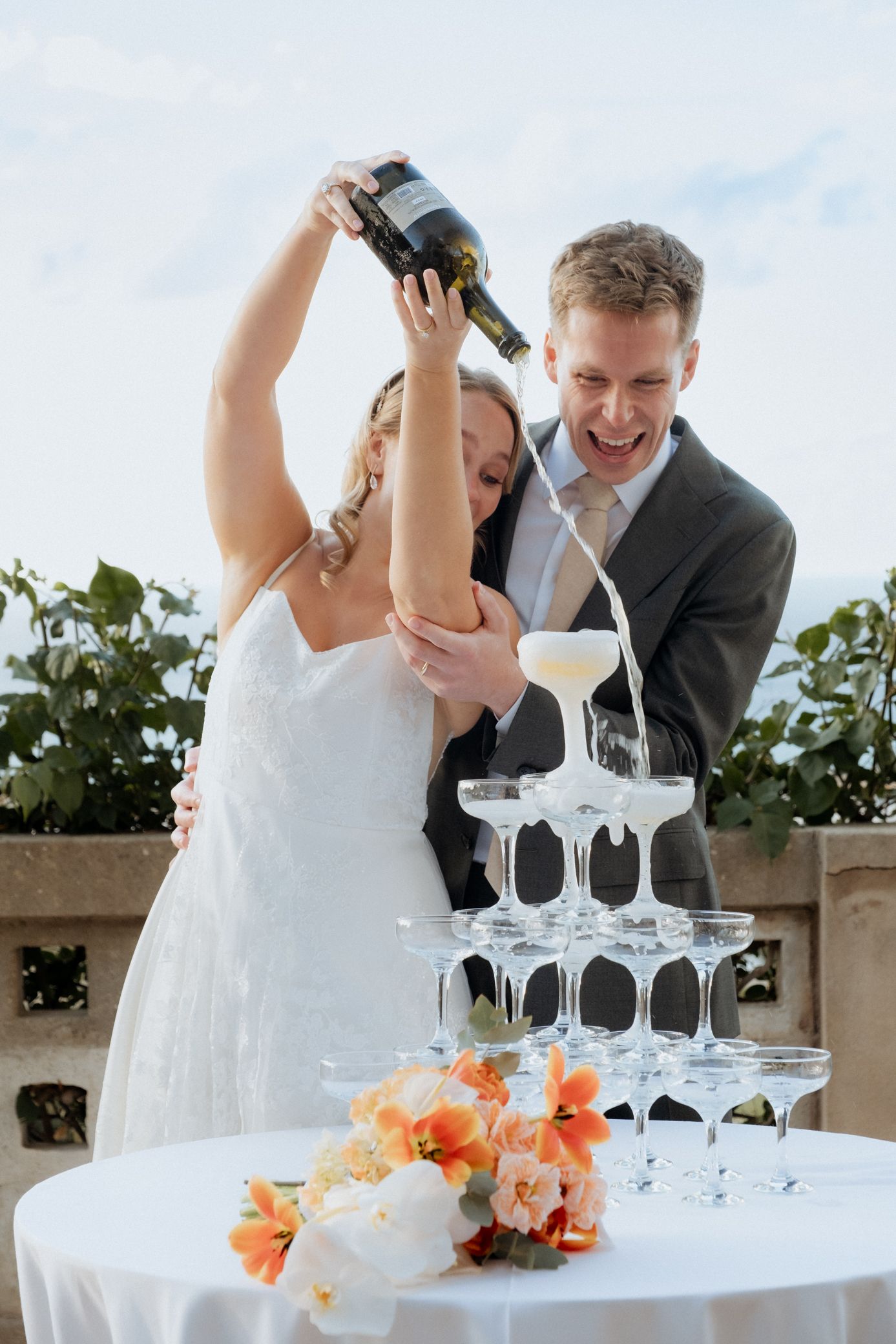 A newlywed couple pours champagne into a tower of crystal glasses during their wedding celebration.