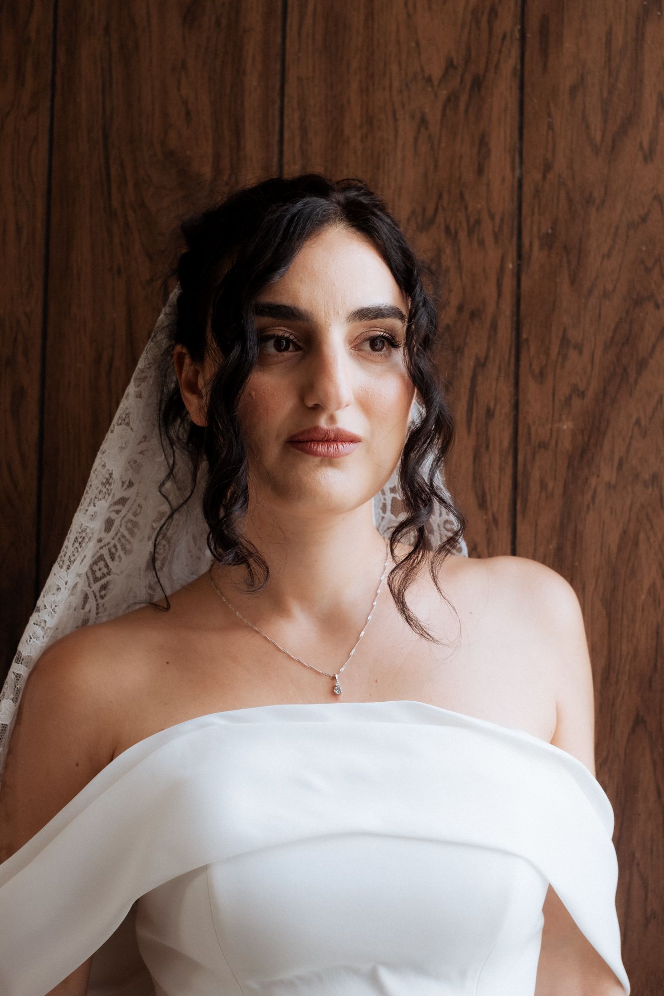 Bride wearing an elegant off-shoulder white wedding gown and lace veil against wooden backdrop.