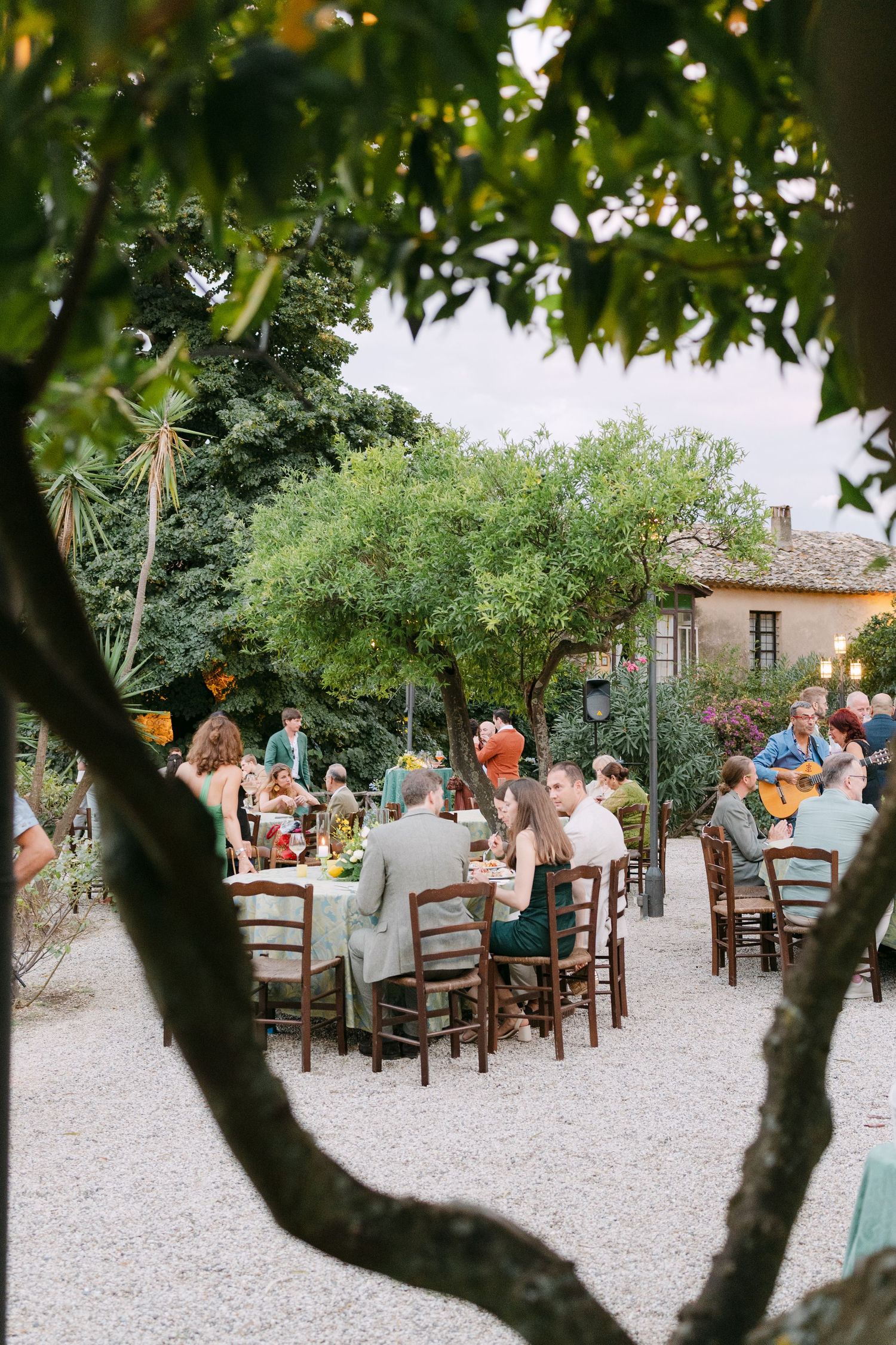 Outdoor wedding reception under tree canopy with guests seated at long tables on a gravel patio.