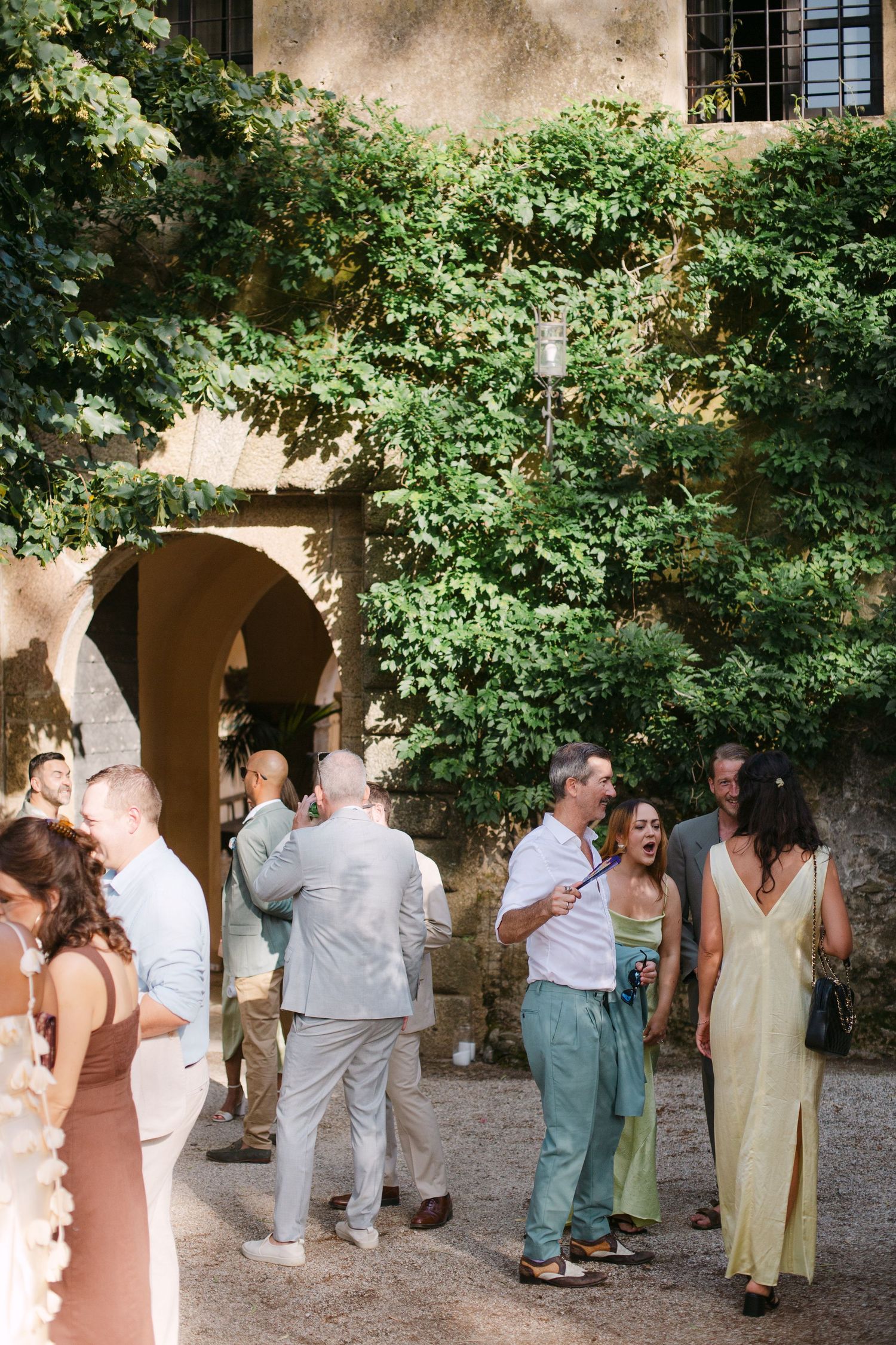 Guests mingle at an outdoor wedding reception near an ivy-covered stone building.
